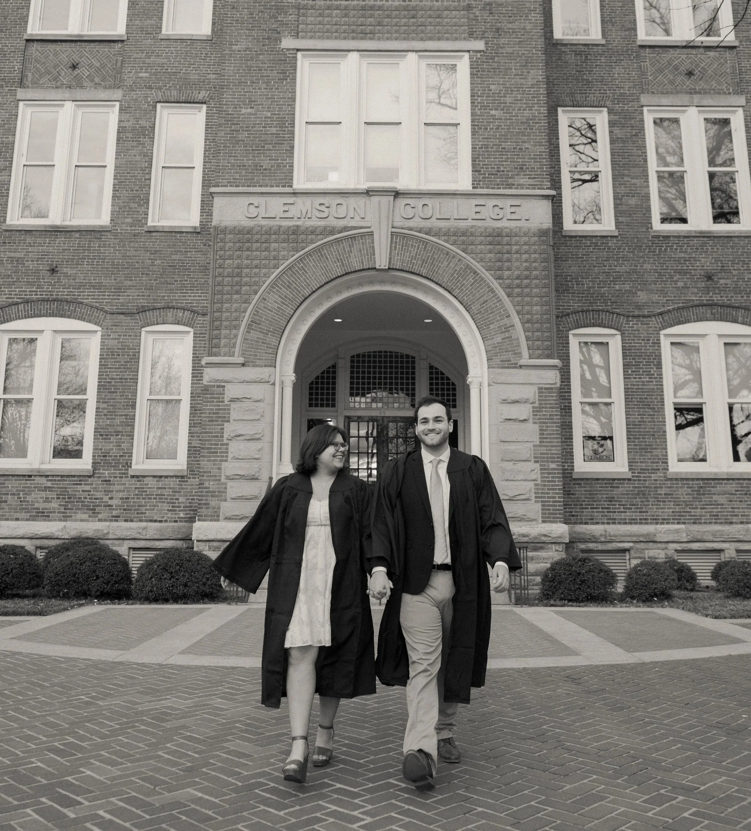 A smiling couple in graduation robes holding hands in front of Clemson College building with brick facade and arched entrance.