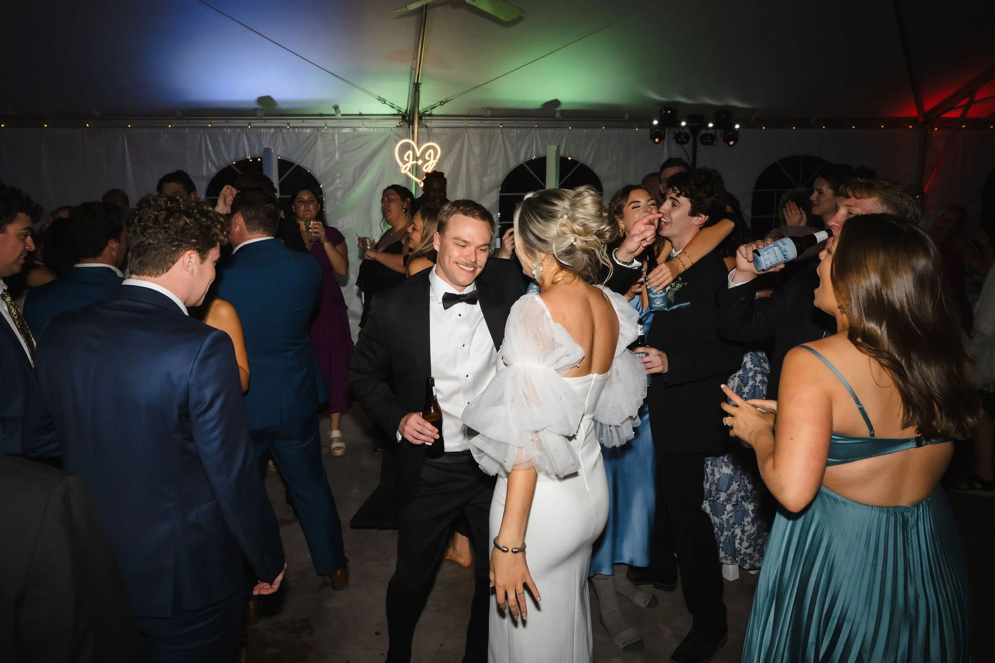 People dancing and socializing at a wedding reception inside a large tent with colorful lighting and a neon 'J&J' sign in the background.