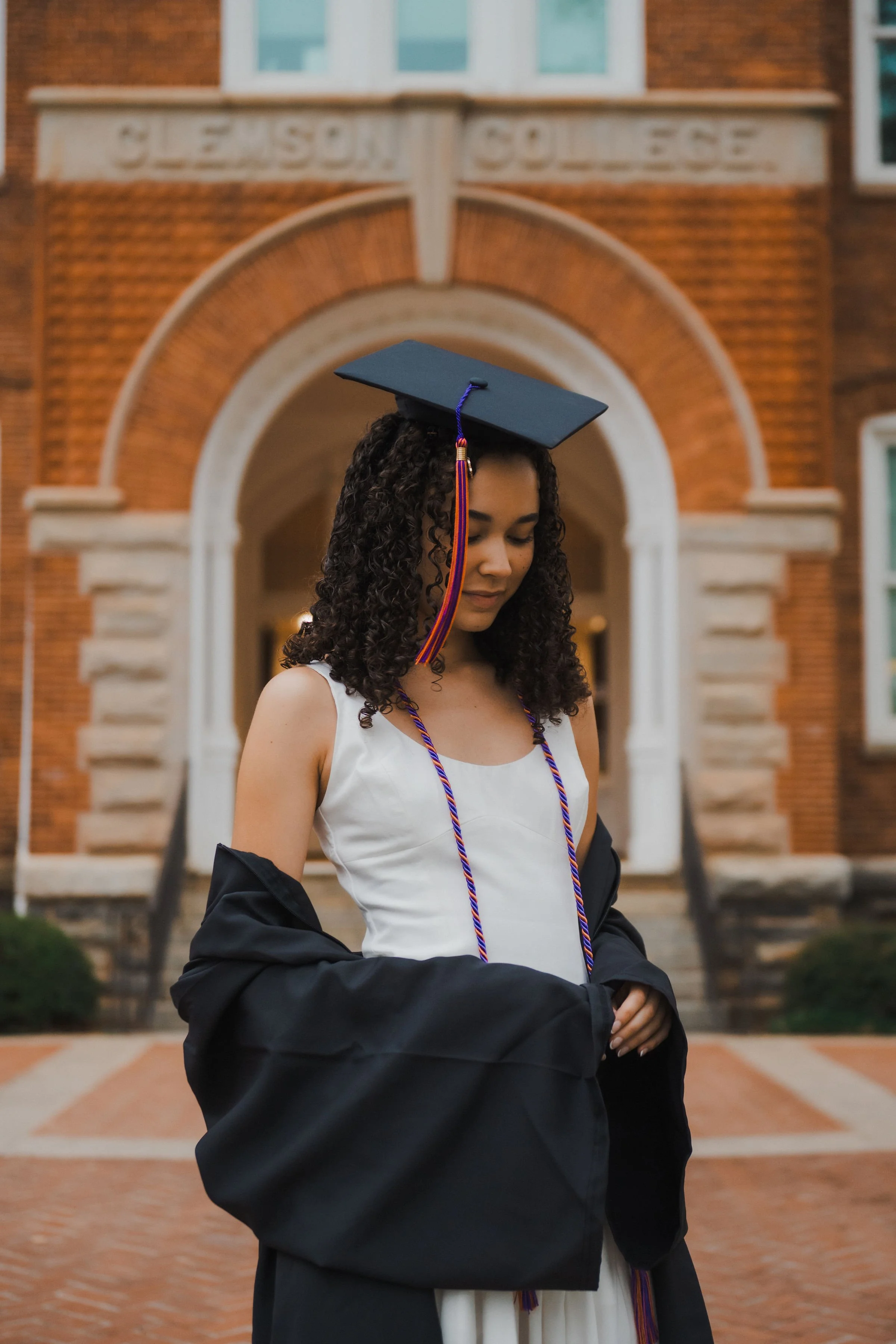 A young woman in a white dress and black graduation gown, with a cap and tassel, stands in front of Clemson College building with brick facade and stone accents.