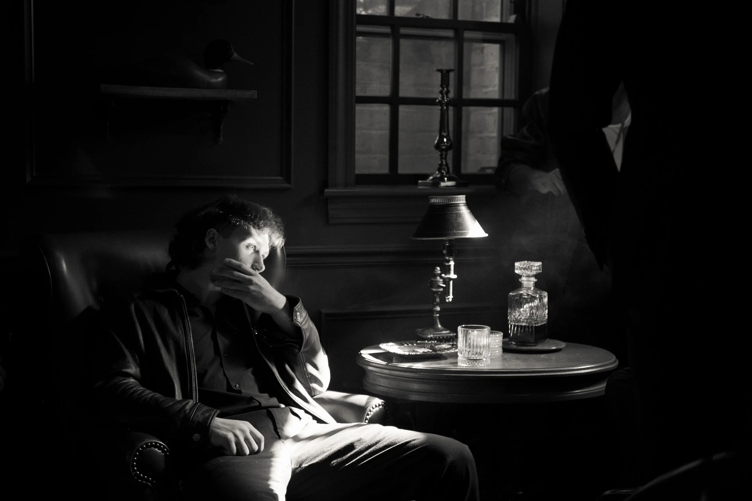 A young man sitting in a dark room, illuminated by a table lamp, with a window behind him. He is wearing a leather jacket and light-colored pants, resting his head on his hand. There is a bottle and glasses on the table, and a person standing near the window on the right side, partially visible.