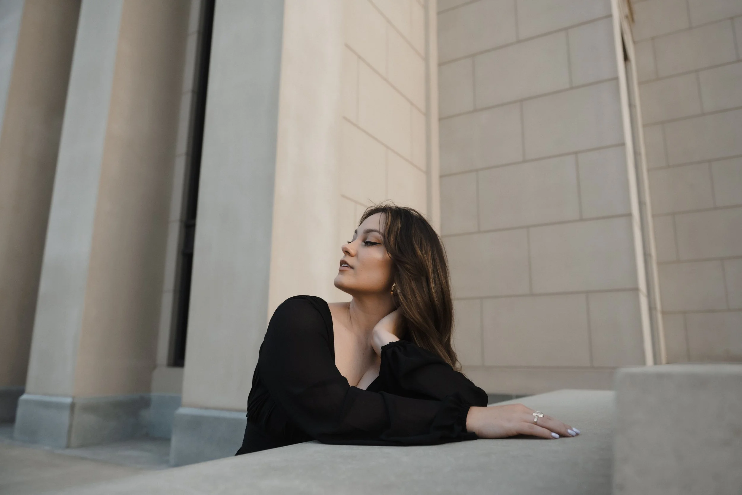 A young woman with long brown hair wearing a black top, leaning on a stone surface with her eyes closed and hand behind her neck, in front of a beige stone building.
