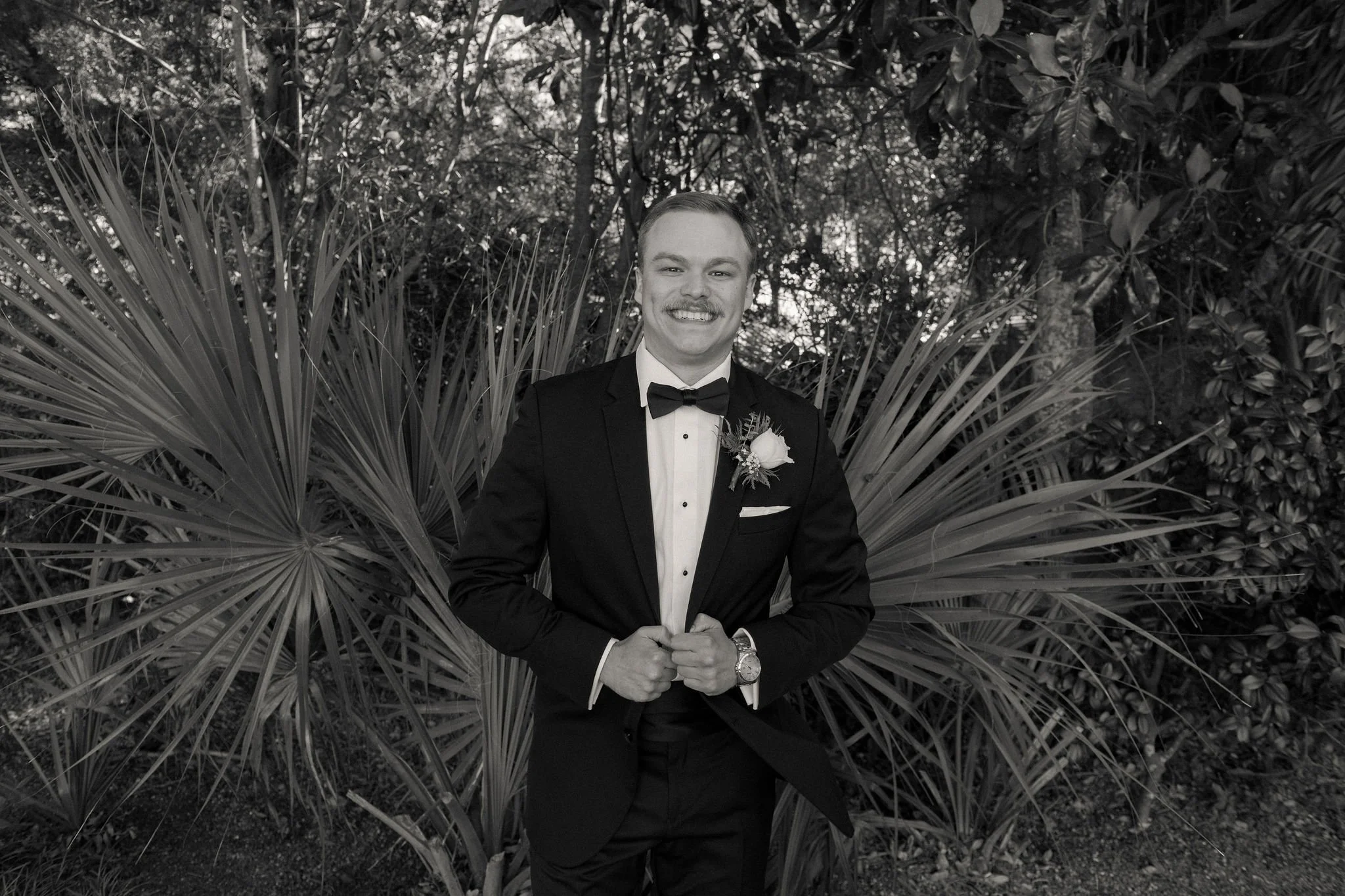 A man dressed in a black tuxedo with a white shirt and black bow tie, standing outdoors in front of large palm leaves and foliage, smiling with hands clasped at waist. He has a boutonniere on his blazer and a watch on his left wrist.