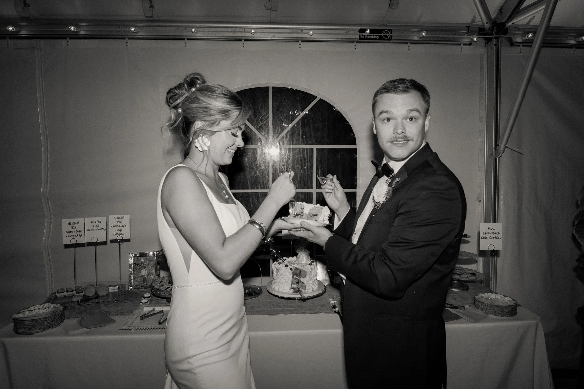 A bride and groom at their wedding reception, sharing a piece of cake. The bride is smiling with her eyes closed, and the groom is looking at the camera. There is a wedding cake on the table behind them, decorated with flowers and candles.