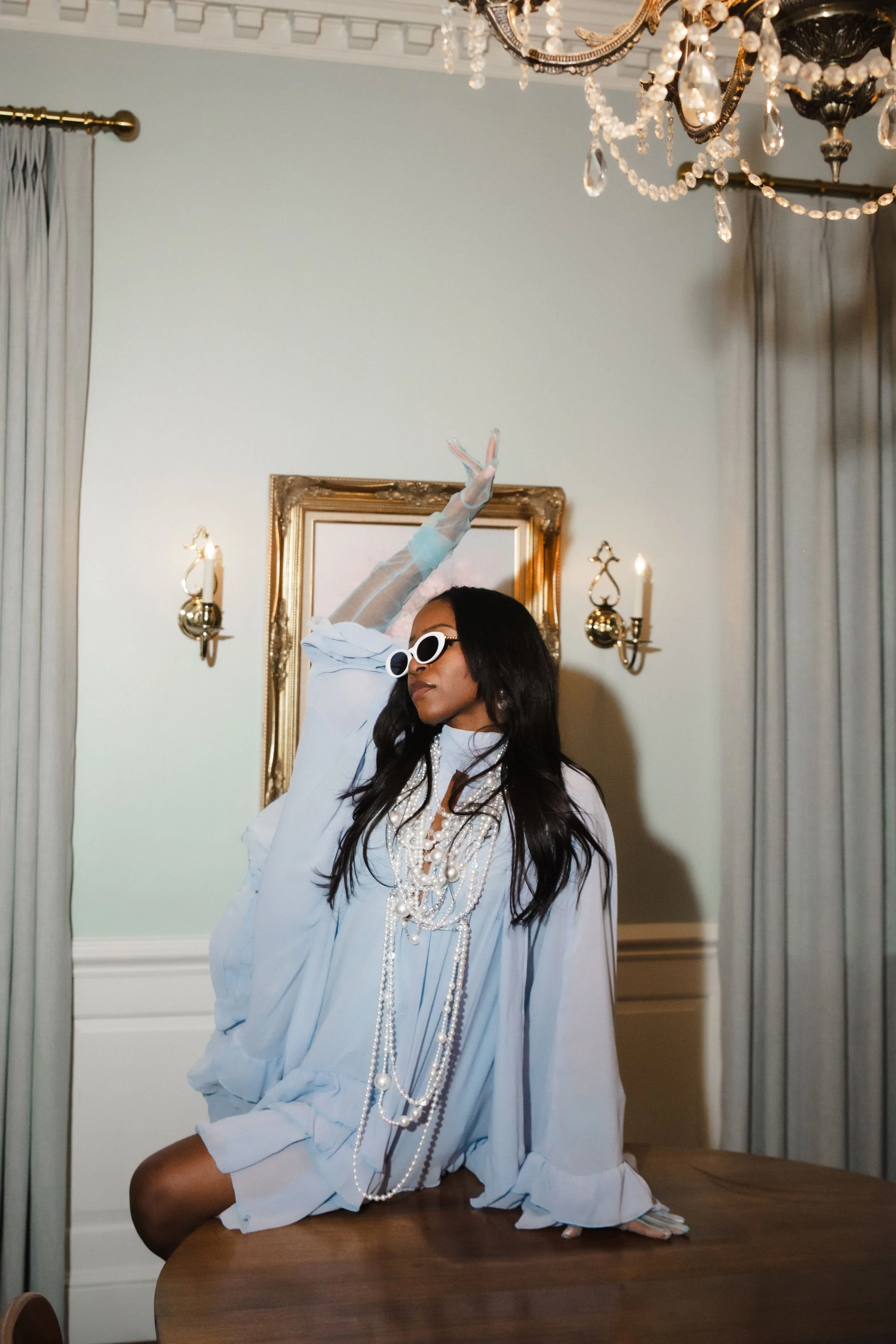 A woman dressed in a white outfit and wearing multiple strands of pearl necklaces. She is sitting on a wooden table in a room with elegant drapes, a large ornate framed mirror, and a chandelier. She is wearing white sunglasses and striking a pose wit