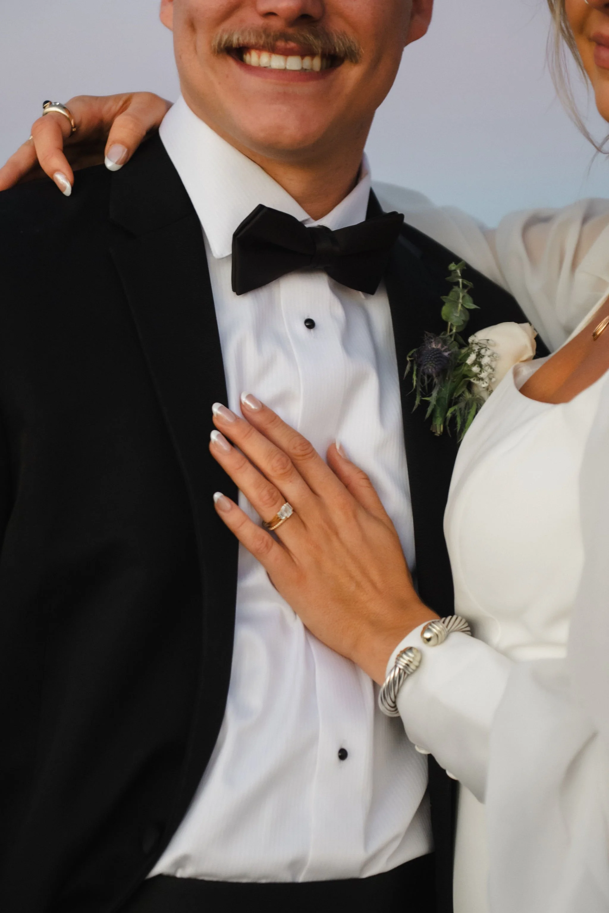 Close-up of a smiling man in a tuxedo with a woman, both dressed formally, with visible wedding rings.