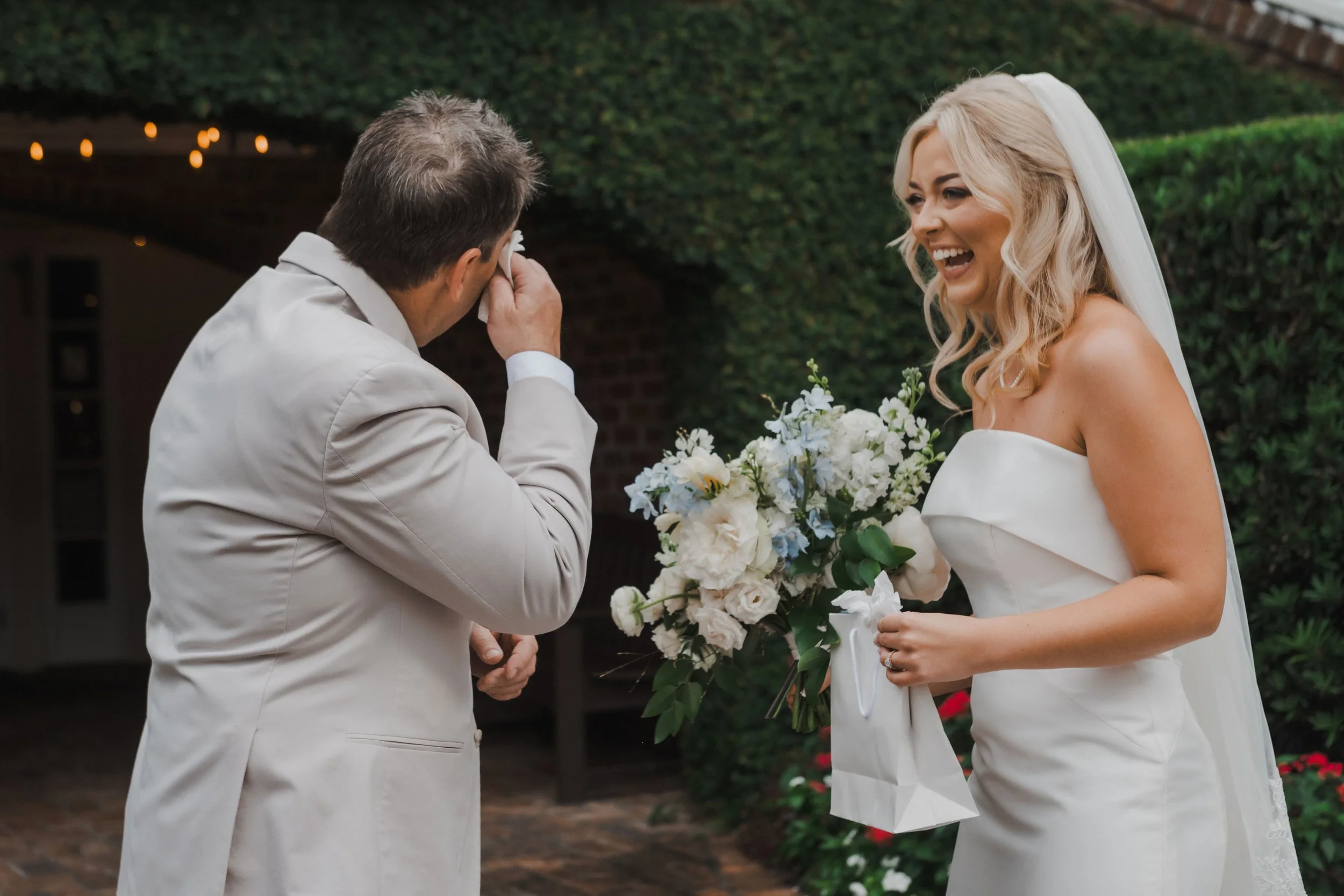A bride with blonde hair in an off-shoulder wedding dress, smiling while holding a bouquet, talking with a man in a beige suit who is wiping his eye, possibly crying, in an outdoor garden setting.