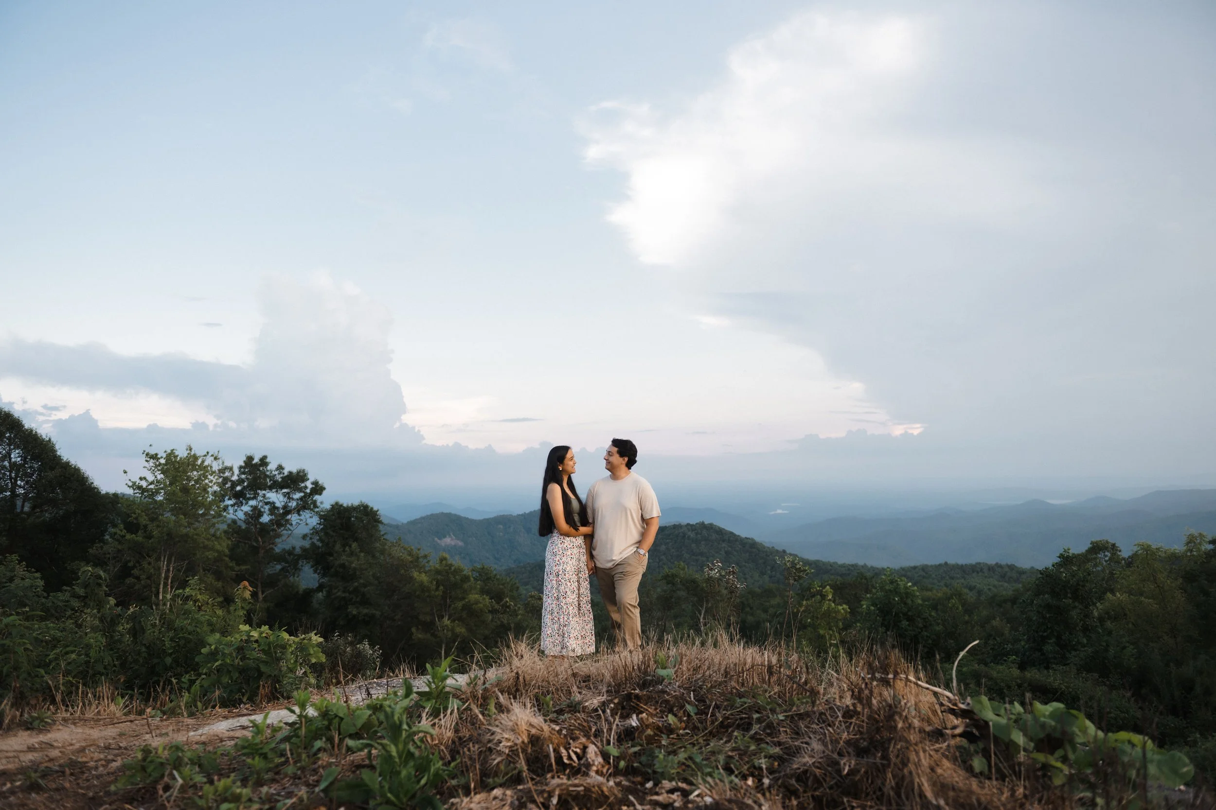A couple standing on a hilltop with scenic mountain and forest landscape at dusk.