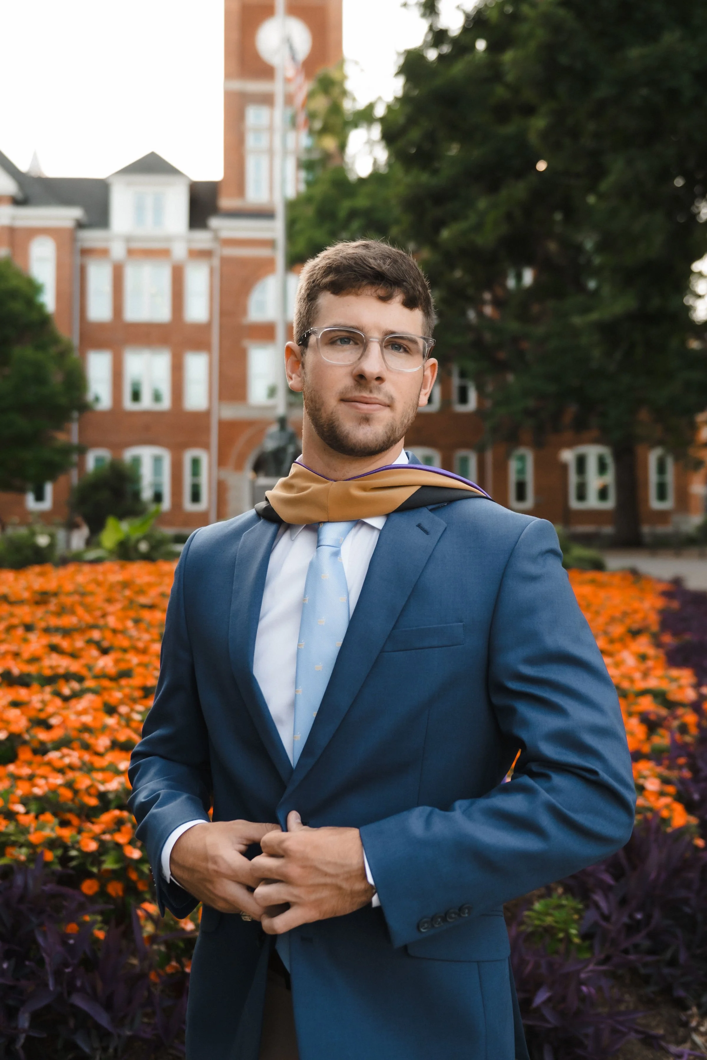 A young man in a blue suit and tie standing outdoors in front of a brick building and flower bed, wearing a graduation gown's hood draped around his shoulders.