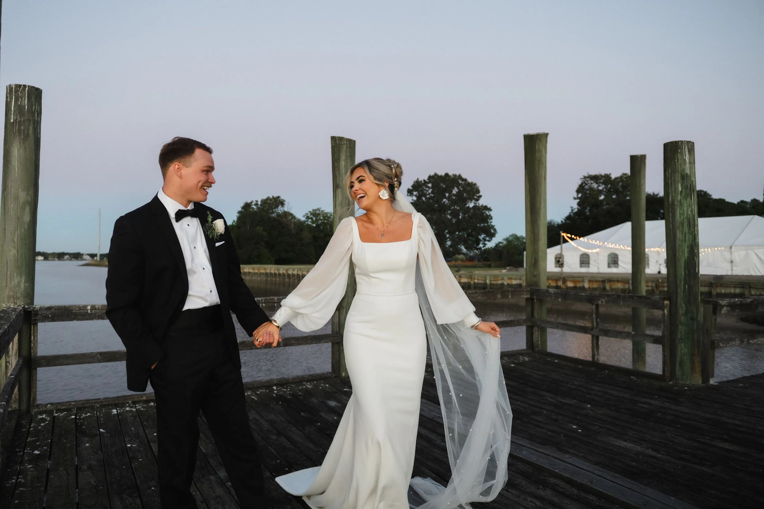 A newlywed couple holding hands and smiling on a wooden dock by the water during sunset, with white tents and string lights in the background.