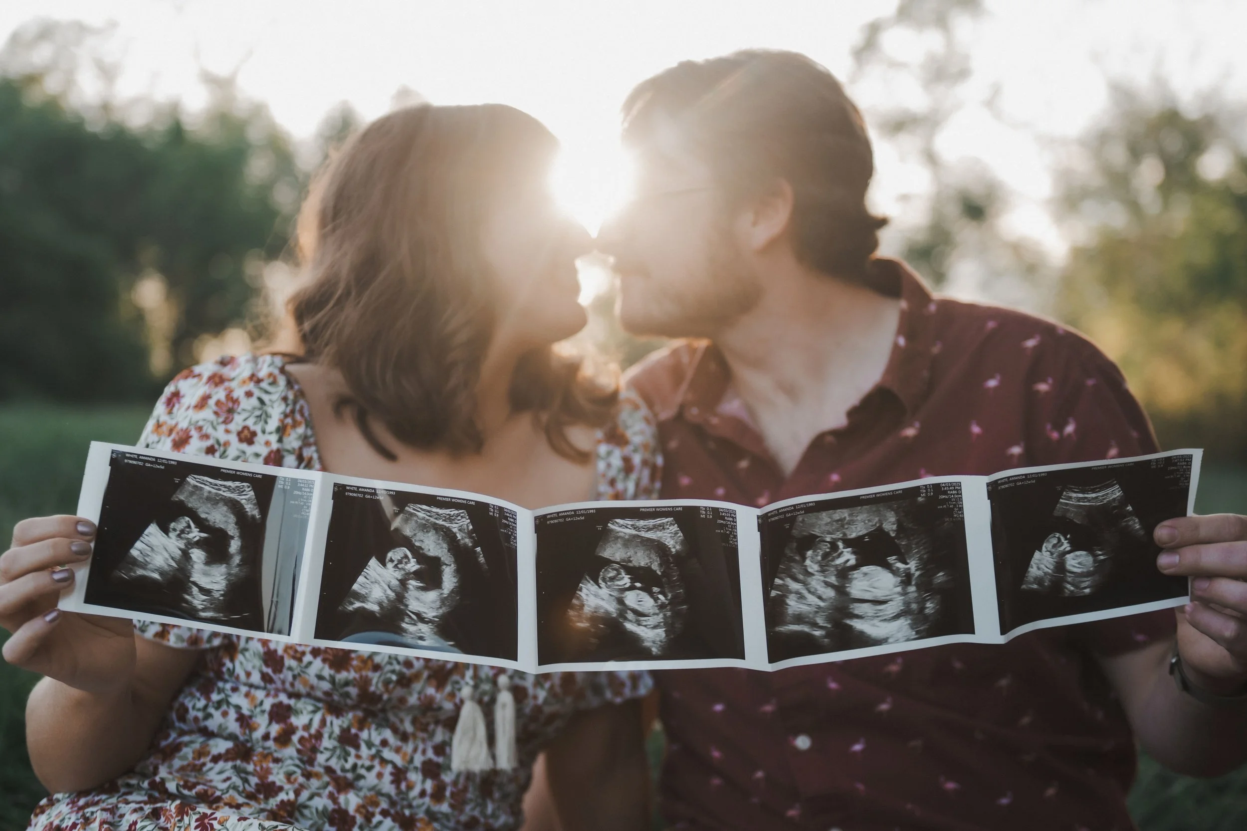 A couple holding ultrasound images of a fetus and touching foreheads at sunset in a park.