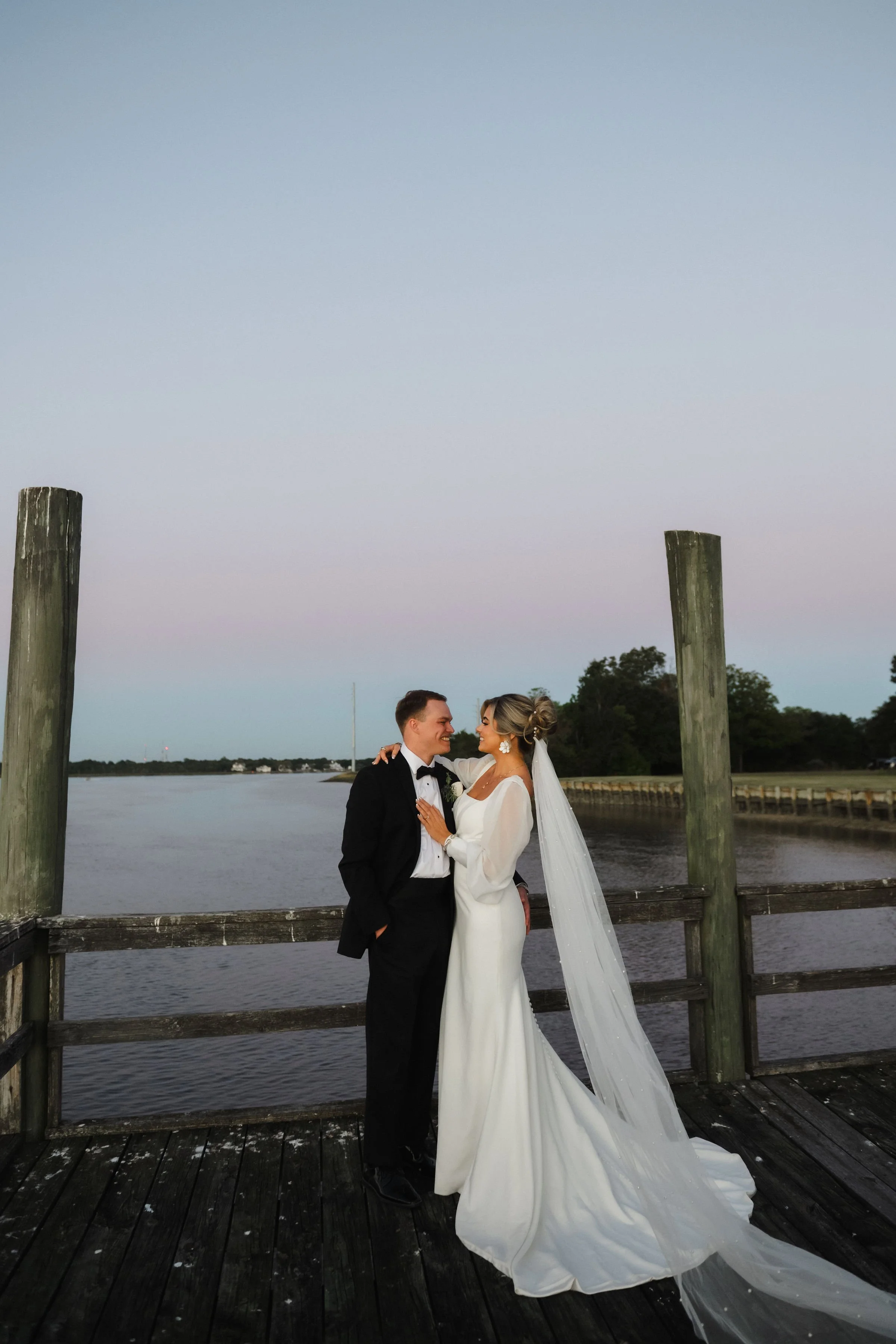 A bride and groom in wedding attire standing on a wooden dock by a river, smiling and looking at each other during sunset or dusk.