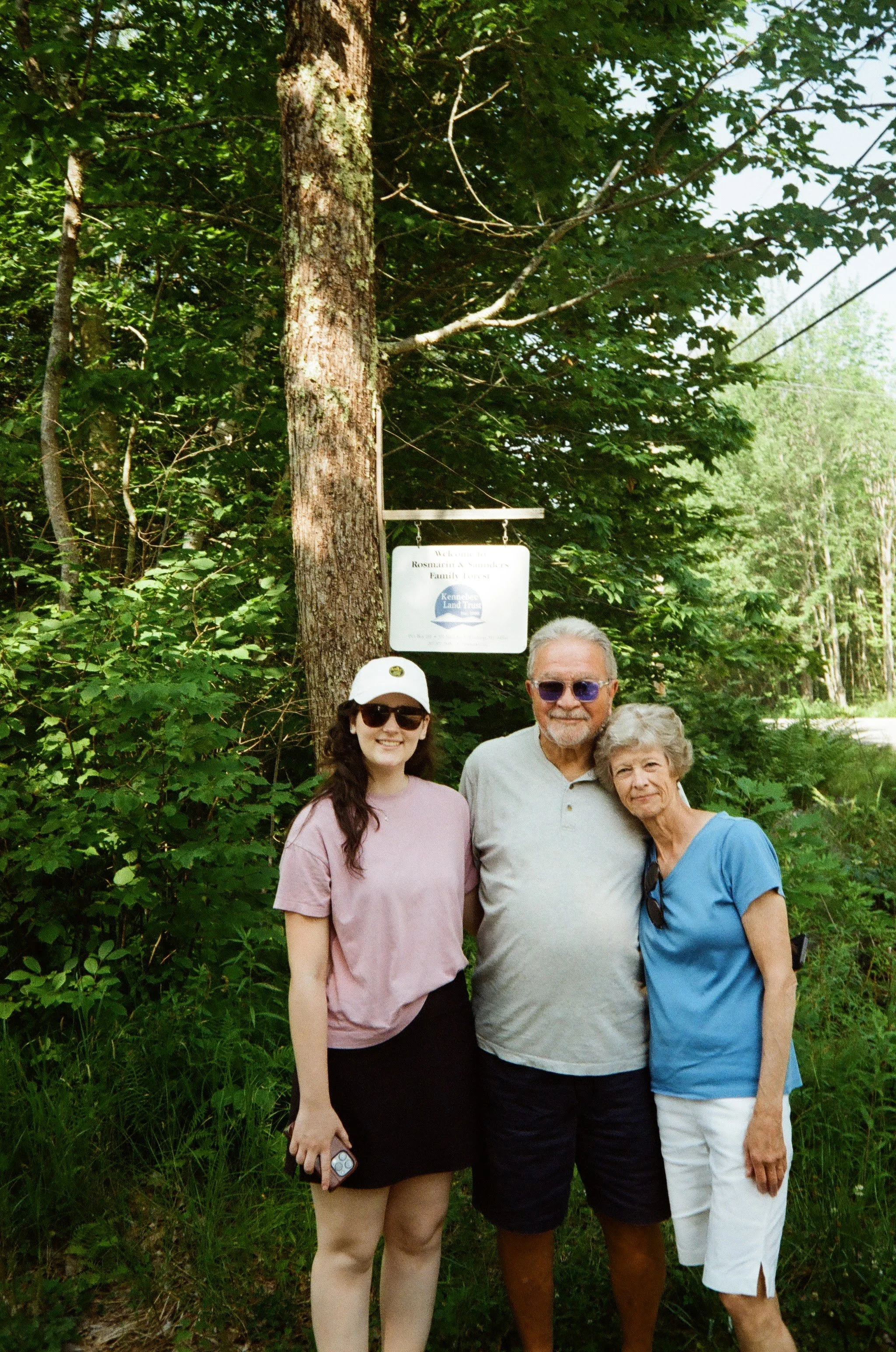 Three people standing outdoors in front of a tree with a sign, smiling at the camera.