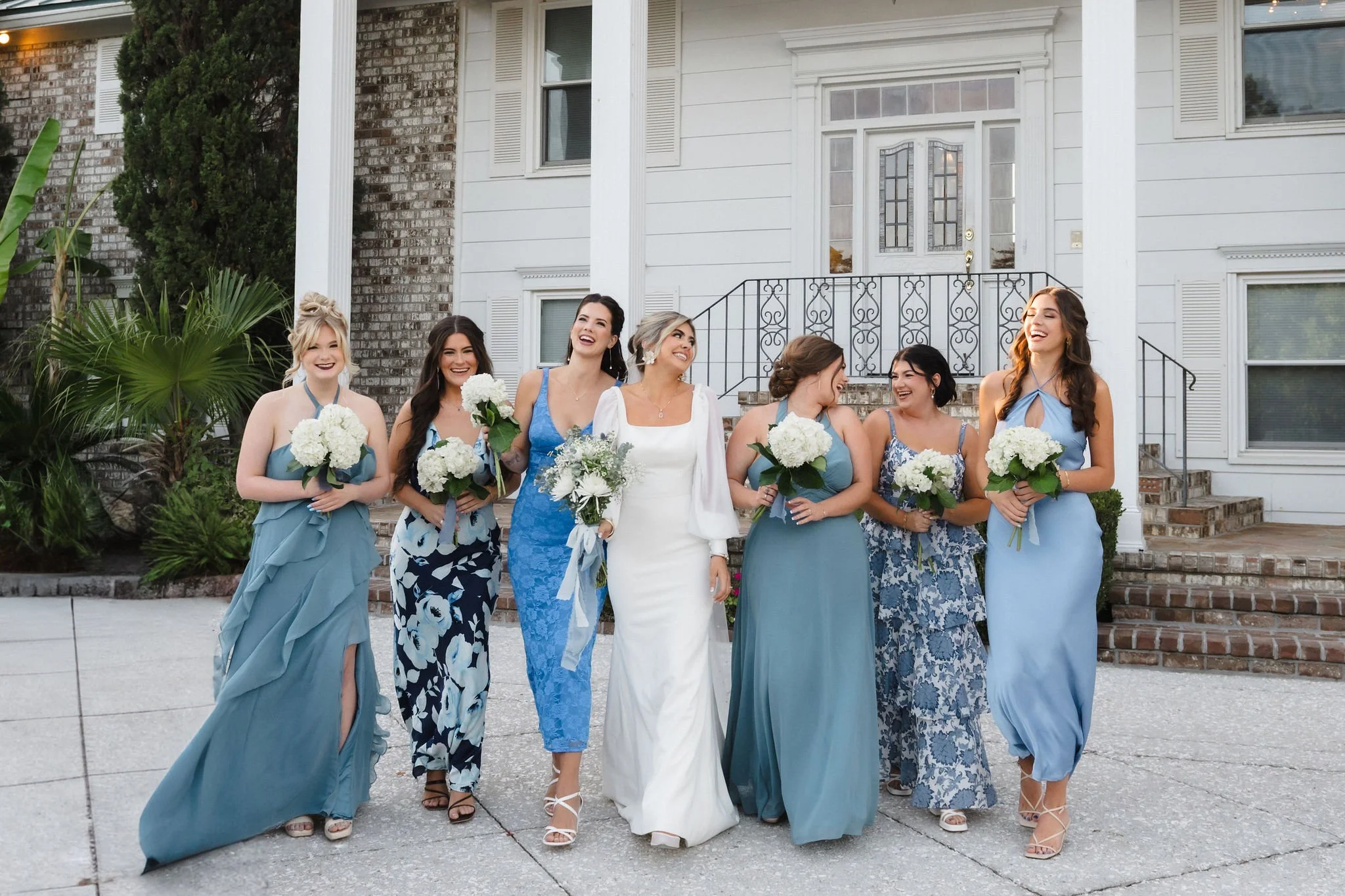 A group of seven women, including a bride in a white dress, walking arm in arm on a sidewalk outside a house, holding bouquets of white flowers, smiling and laughing.