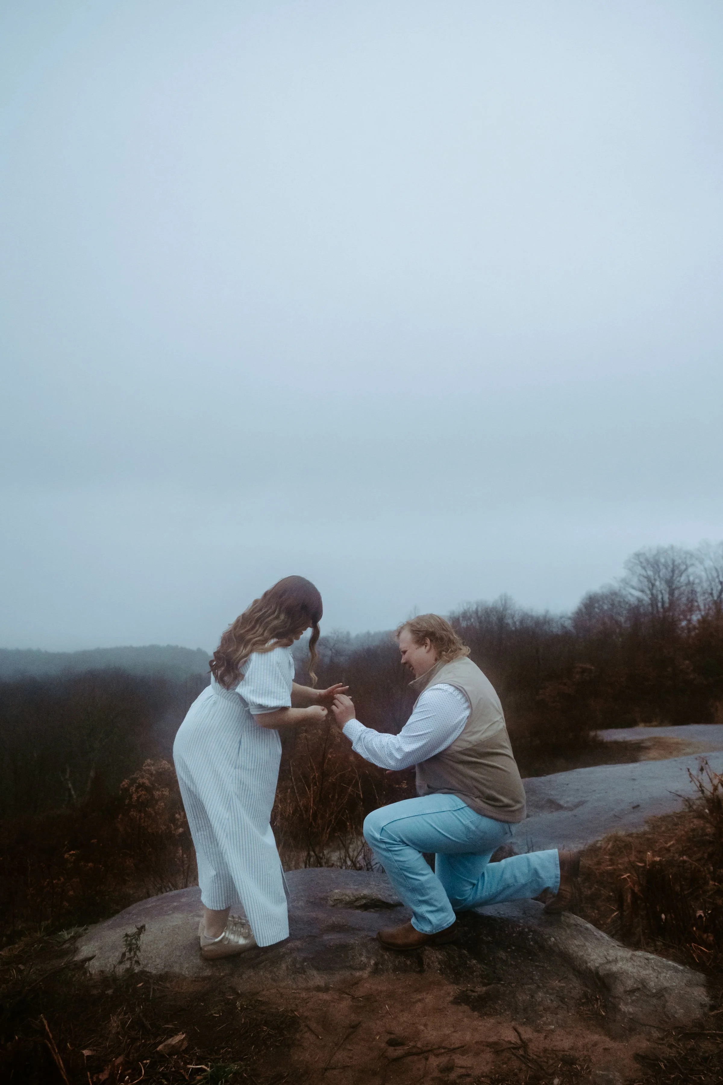 A man on one knee proposing to a woman outdoors on a rocky surface with a cloudy sky and trees in the background.