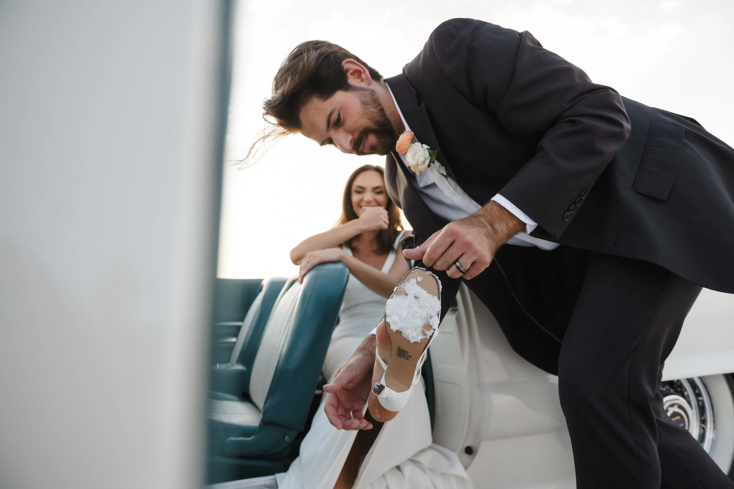 A groom helping his bride with her shoe with a woman in a white dress sitting in the background.