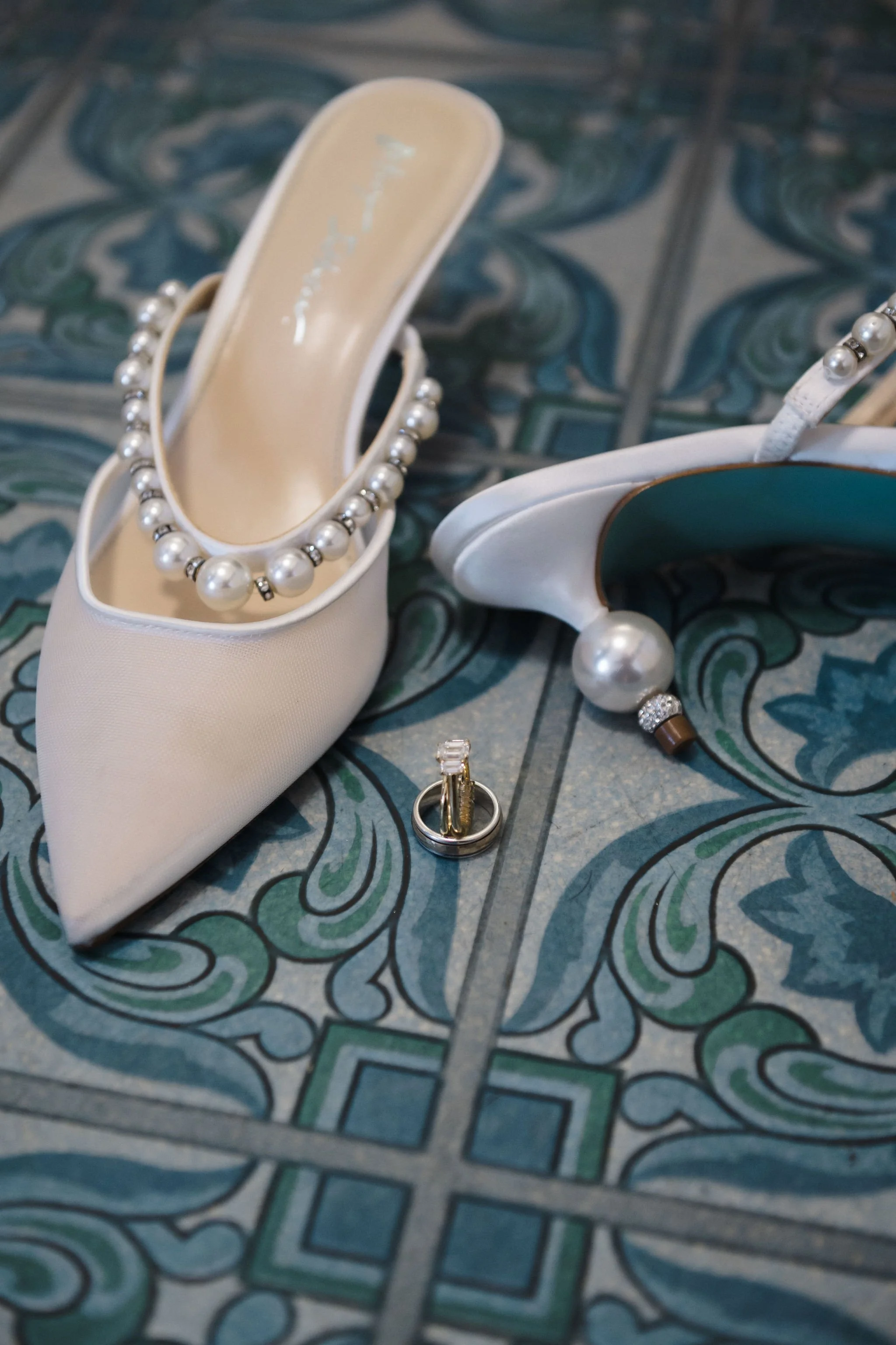 A pair of white high-heeled shoes decorated with pearl straps, two wedding rings, and a pearl bracelet on a decorative tiled floor.
