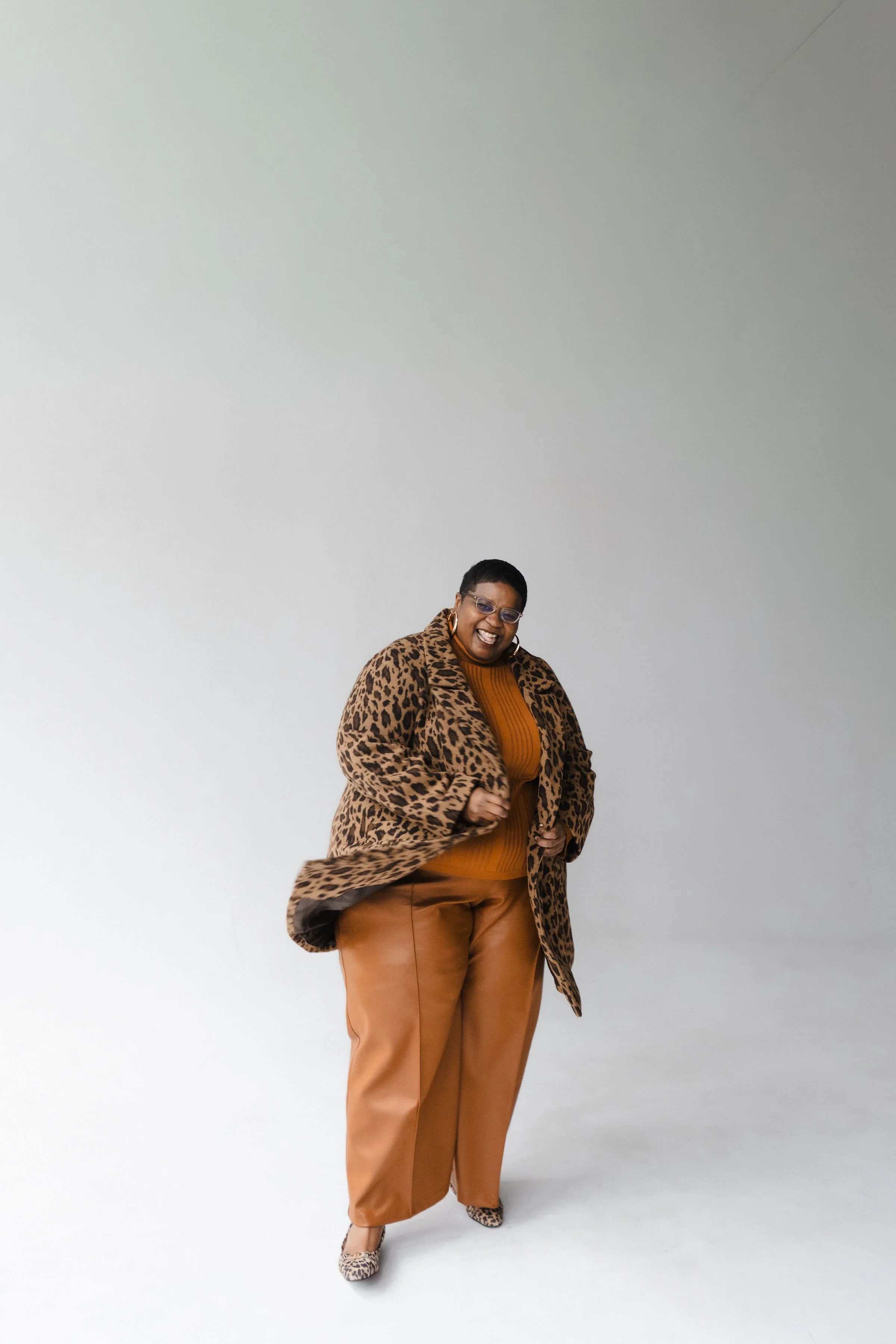 A woman wearing a leopard print coat, mustard sweater, matching leopard print shoes, and brown pants, standing and smiling against a plain white background.