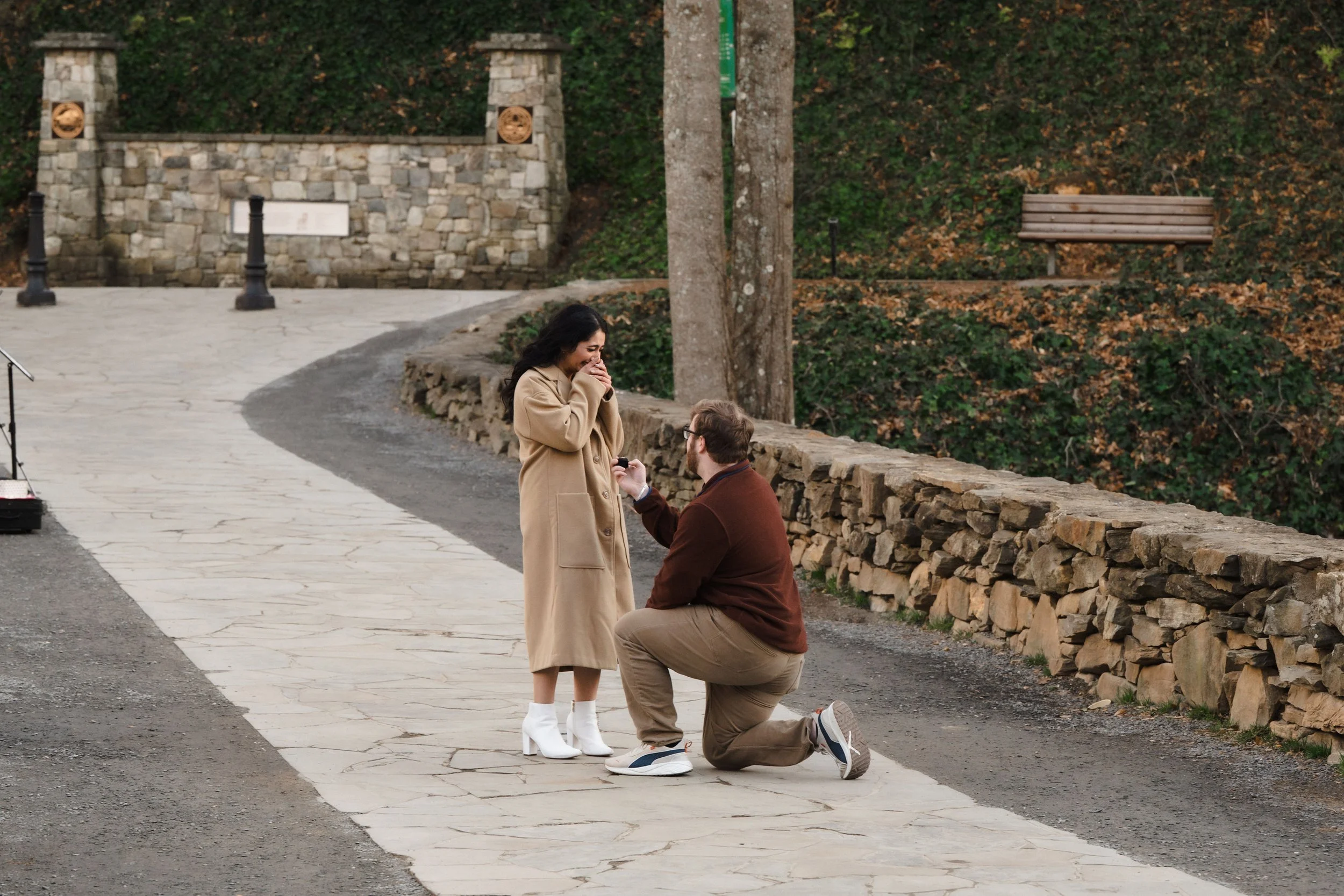 A man on one knee proposing to a woman on a stone-paved pathway in a park, with a scenic background of trees, a stone wall, and benches.