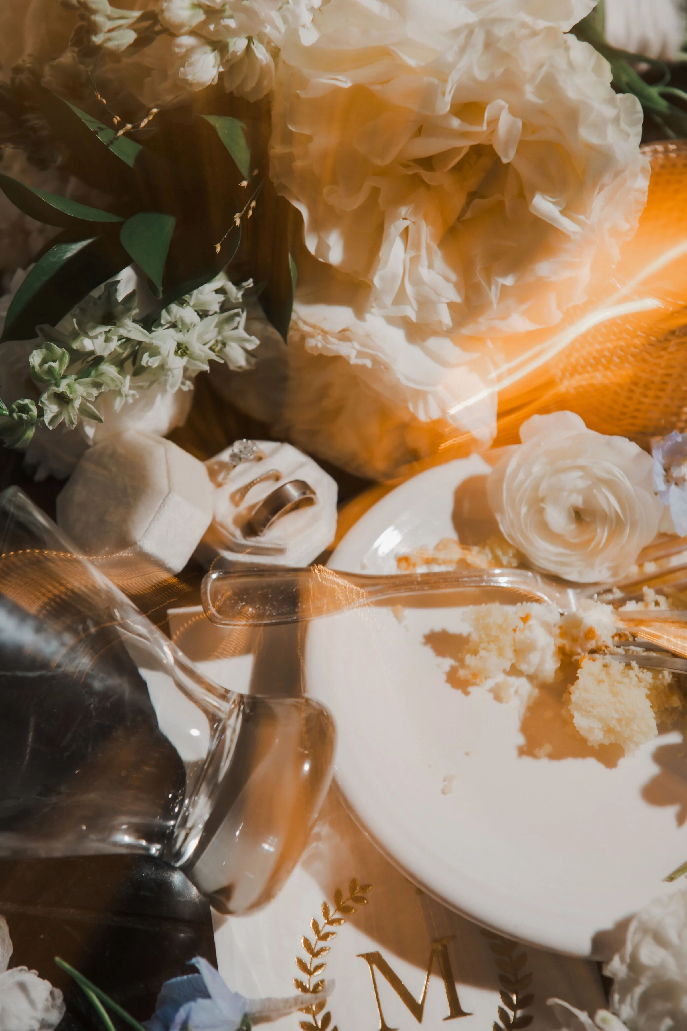 A partially eaten slice of cake on a white plate surrounded by flowers, rings, a glass pitcher, and a decorated letter 'M' with gold laurel accents.