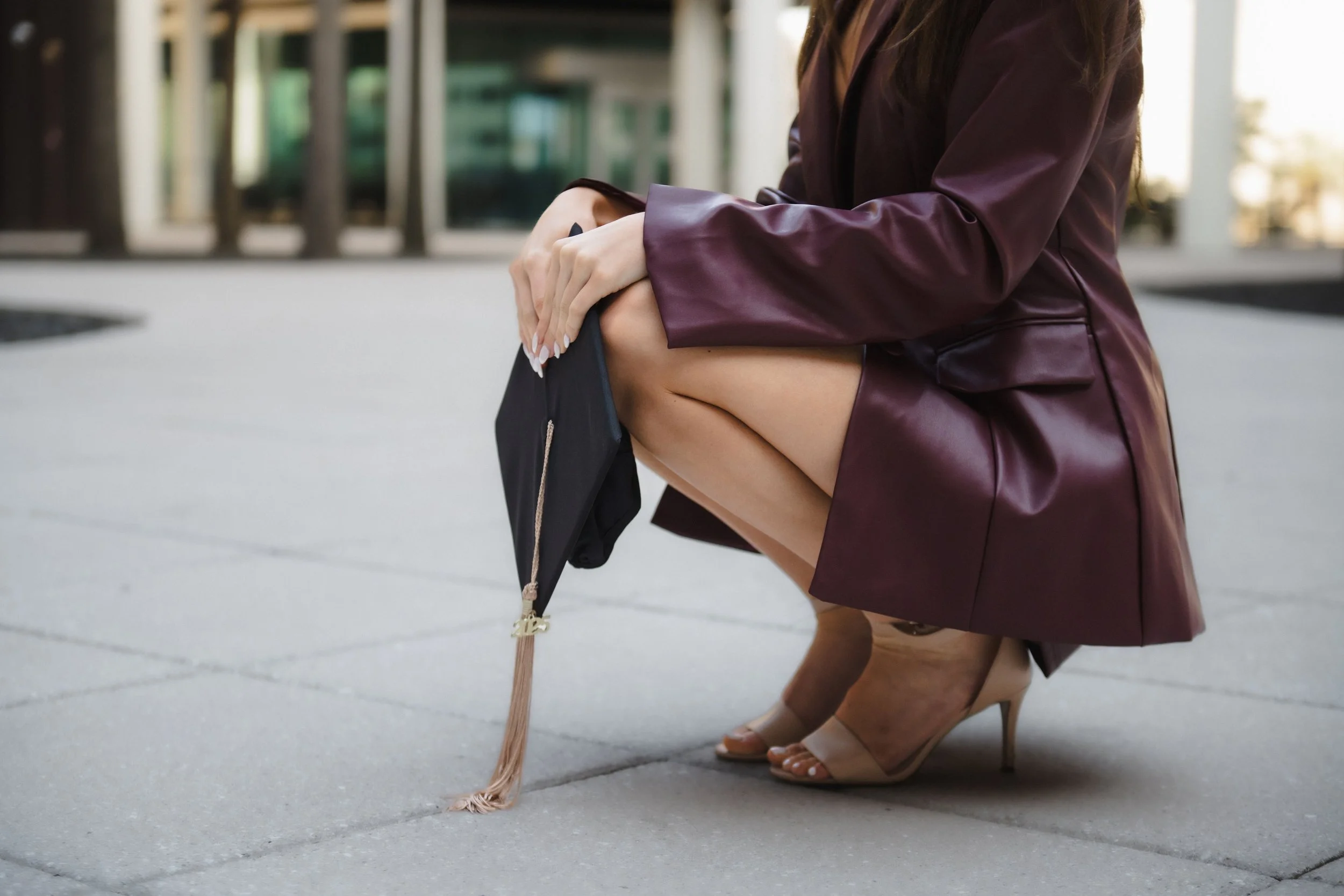 A woman squatting outdoors on a paved surface, holding a black graduation cap with a pink tassel.