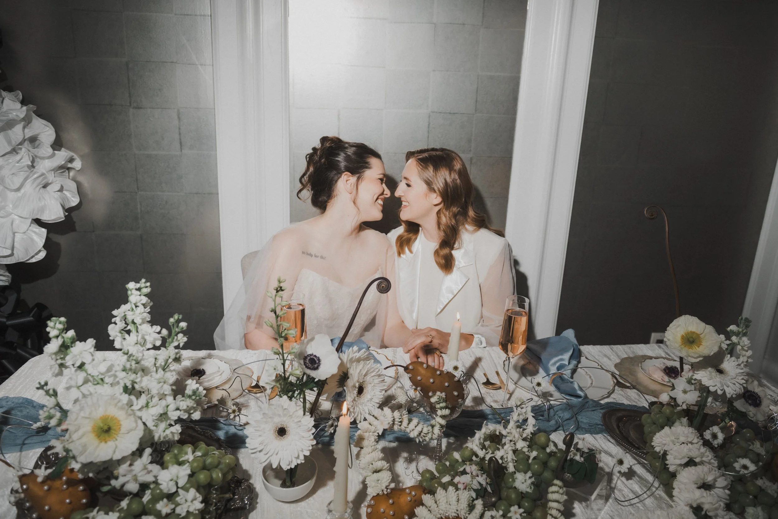 Two women sitting close together at a decorated table, smiling and touching foreheads. One is in a white lace dress and veil, and the other is in a beige blazer.
