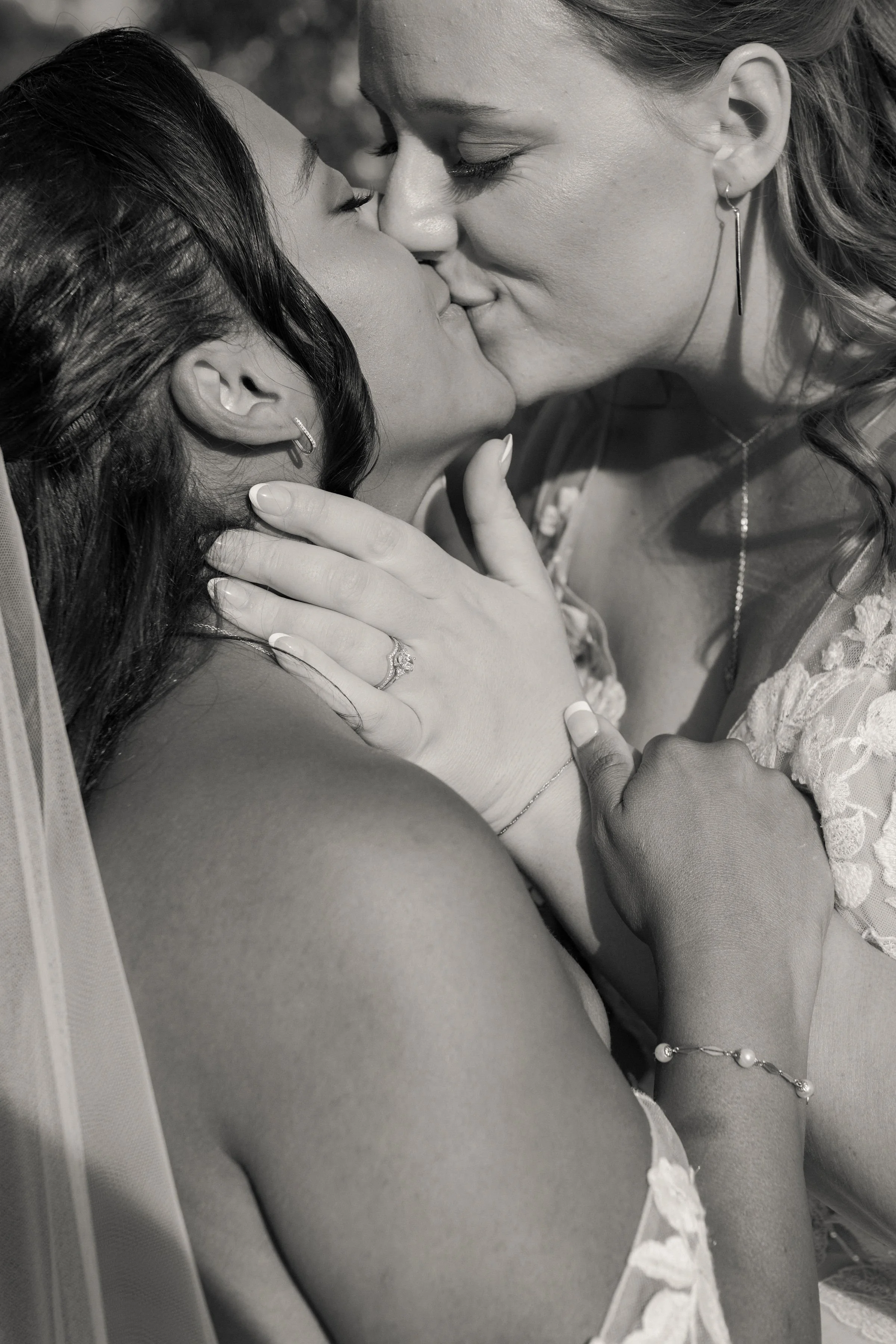Two women sharing a tender kiss, one with dark hair and the other with light hair, both wearing jewelry and wedding rings, in a close intimate pose.