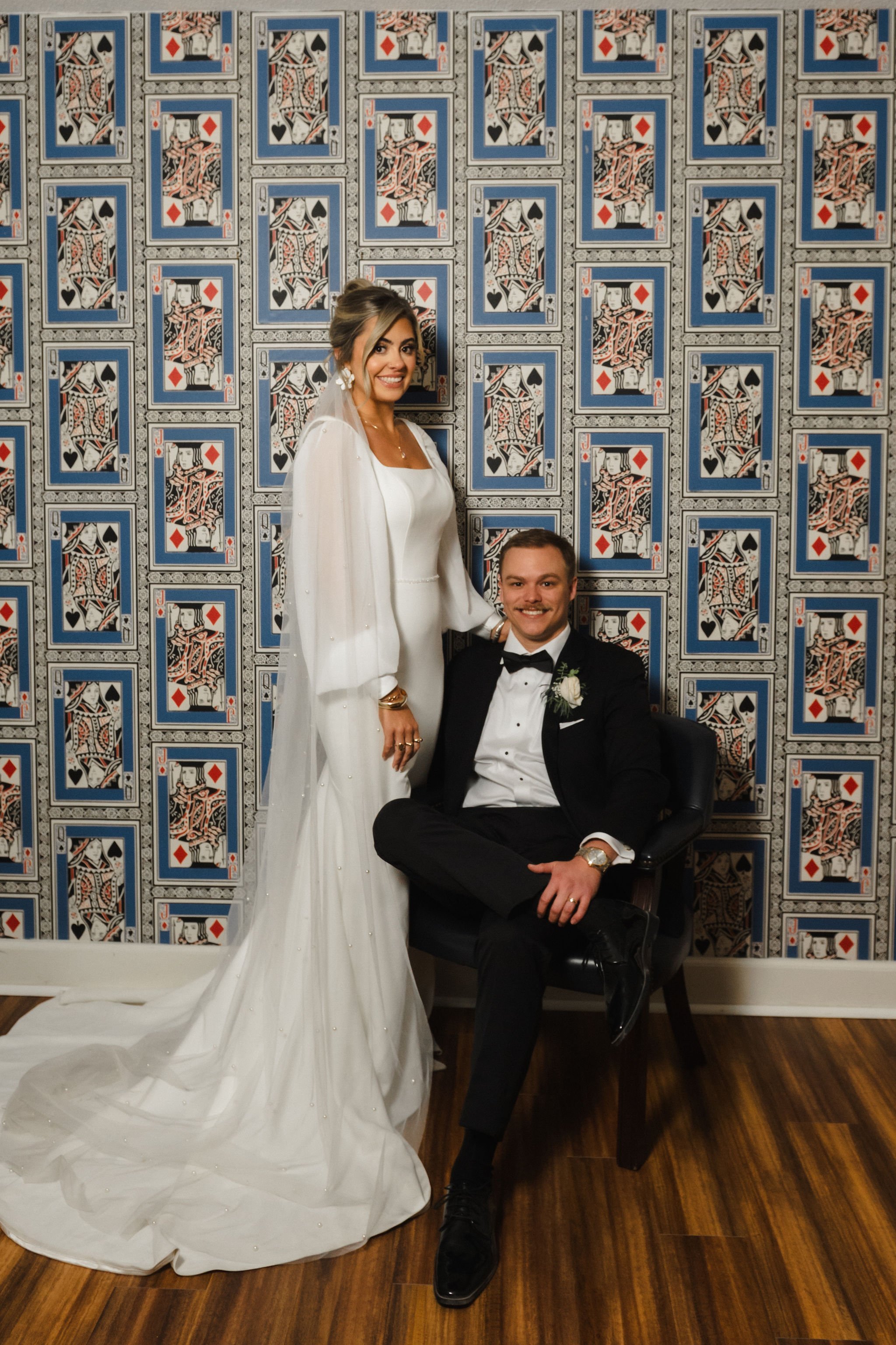 A bride in a white wedding dress and a groom in a black tuxedo sitting against a wall decorated with playing cards patterns.