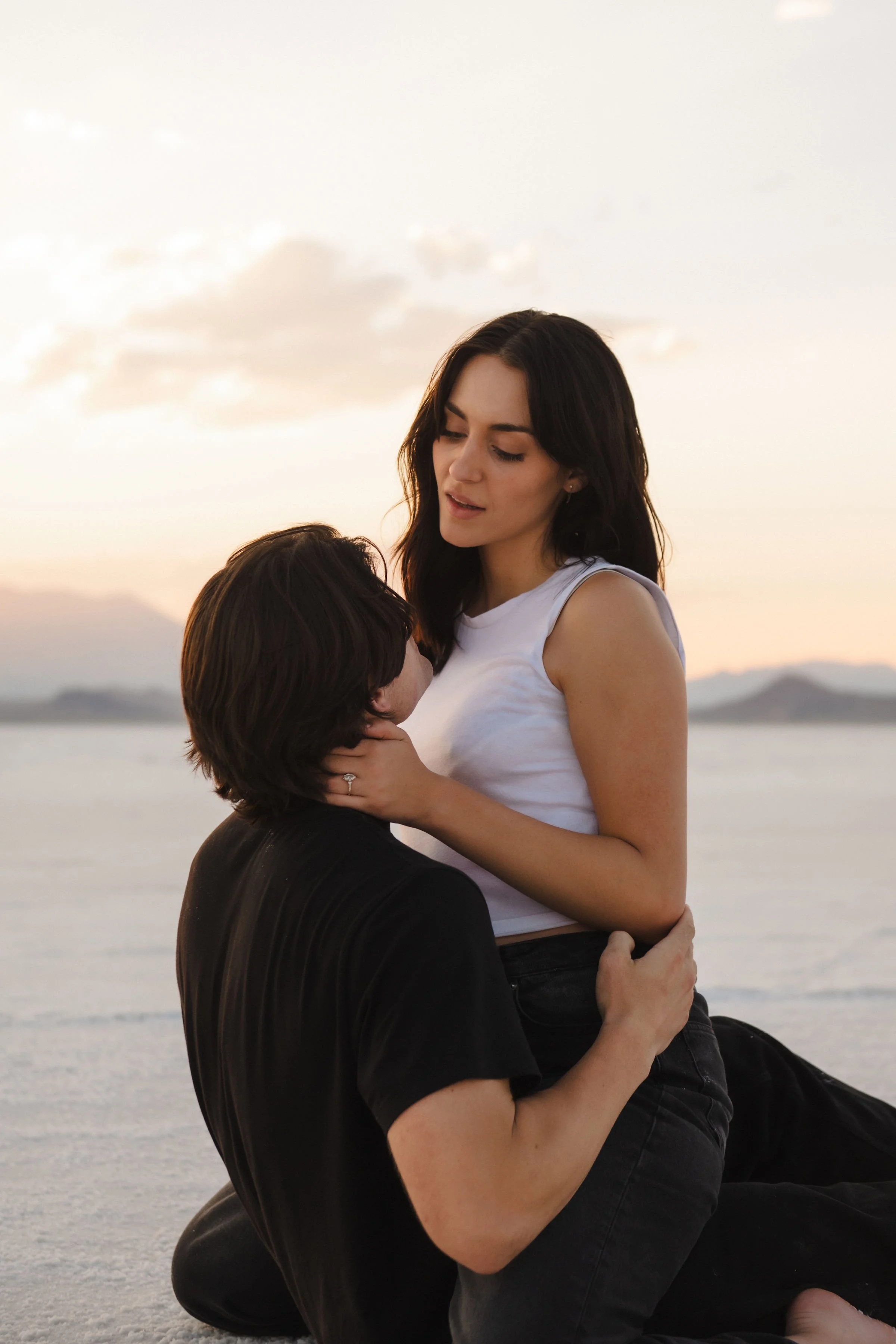 A woman is sitting on the ground by a body of water during sunset, holding a man who is kneeling in front of her, with their faces close together.