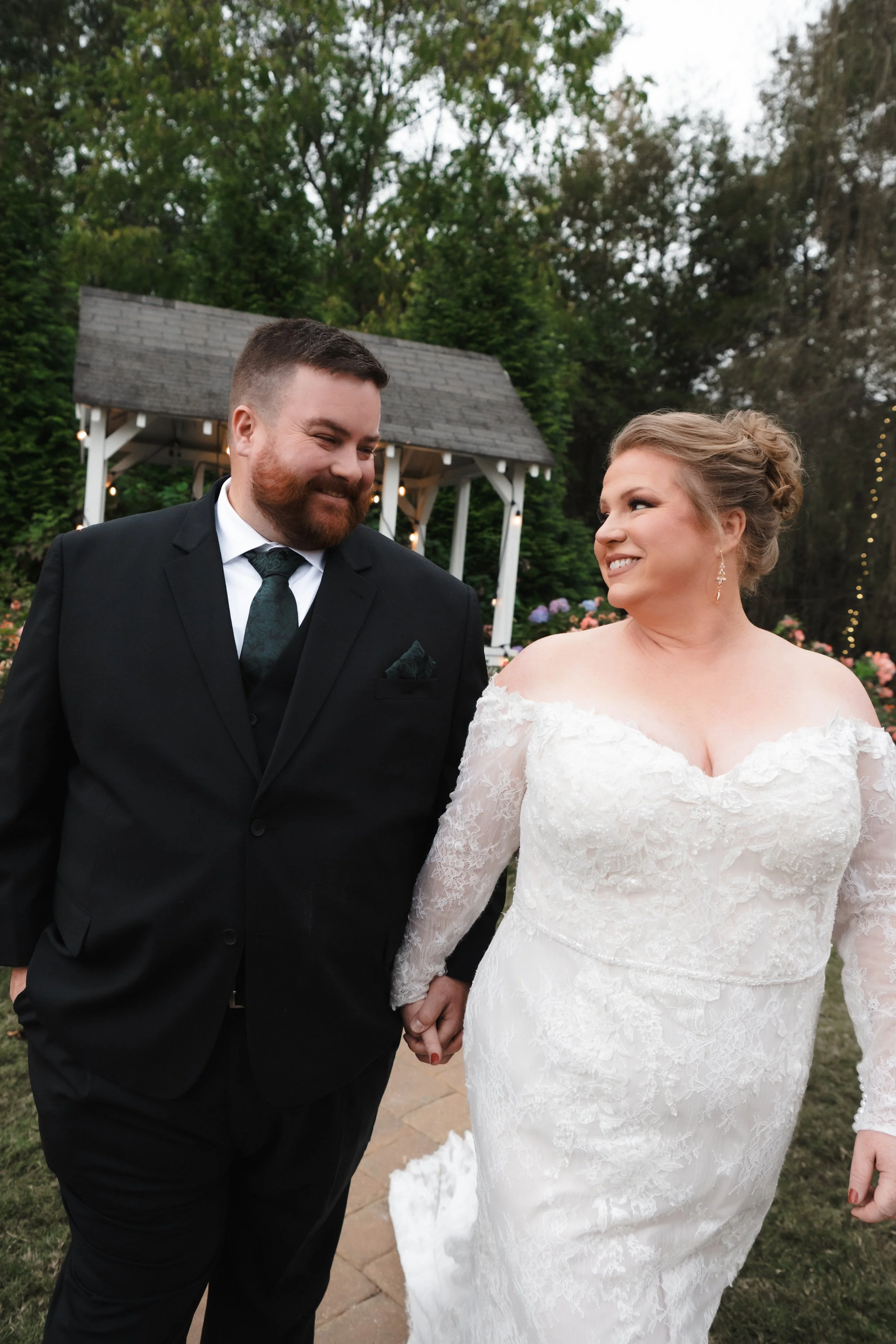 A bride and groom holding hands outdoors, smiling at each other during their wedding ceremony, with a garden and a small white pavilion in the background.