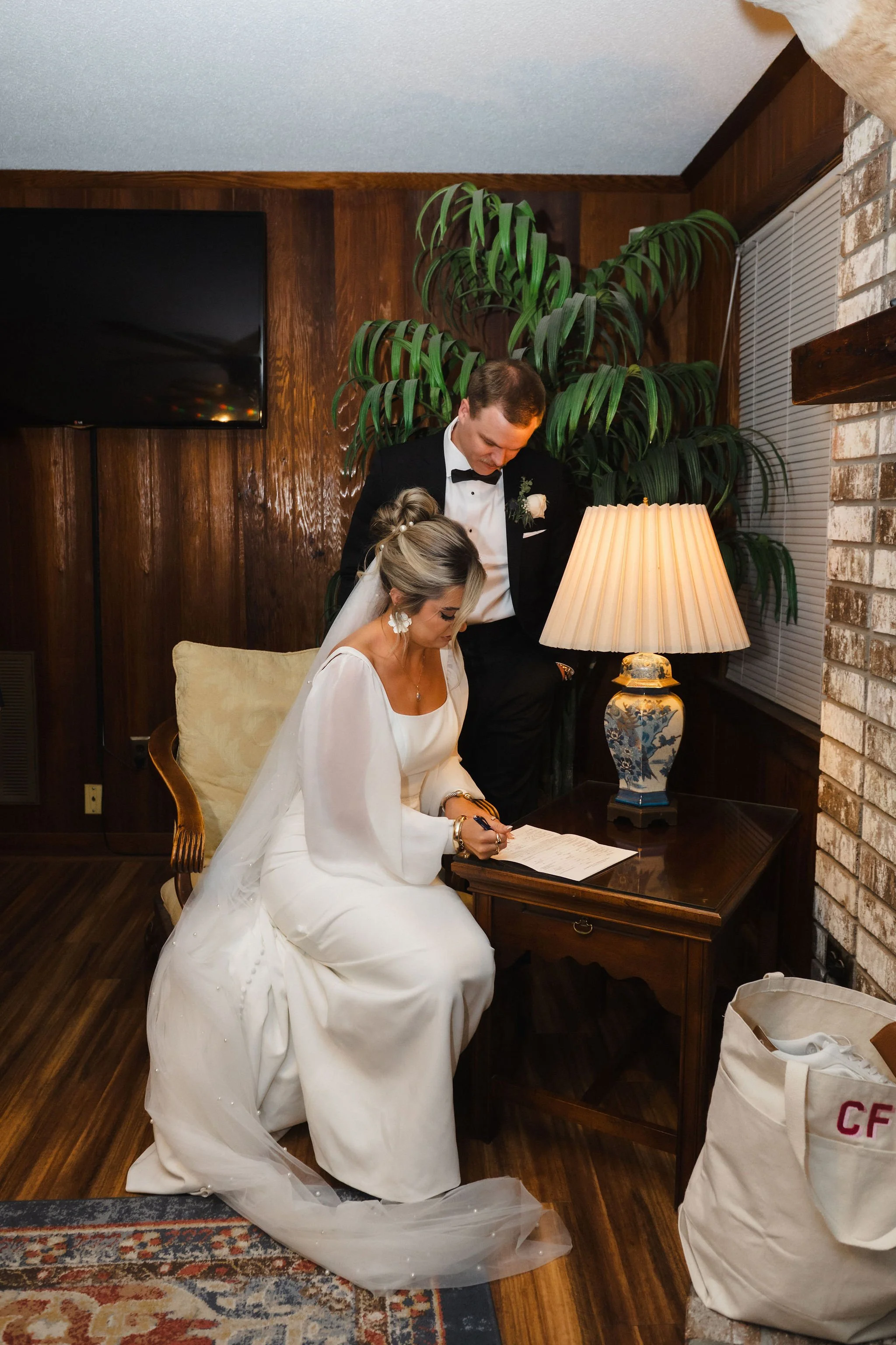 A bride and groom signing a document during their wedding ceremony in a cozy room with wood-paneled walls, a large potted plant, and a table lamp.
