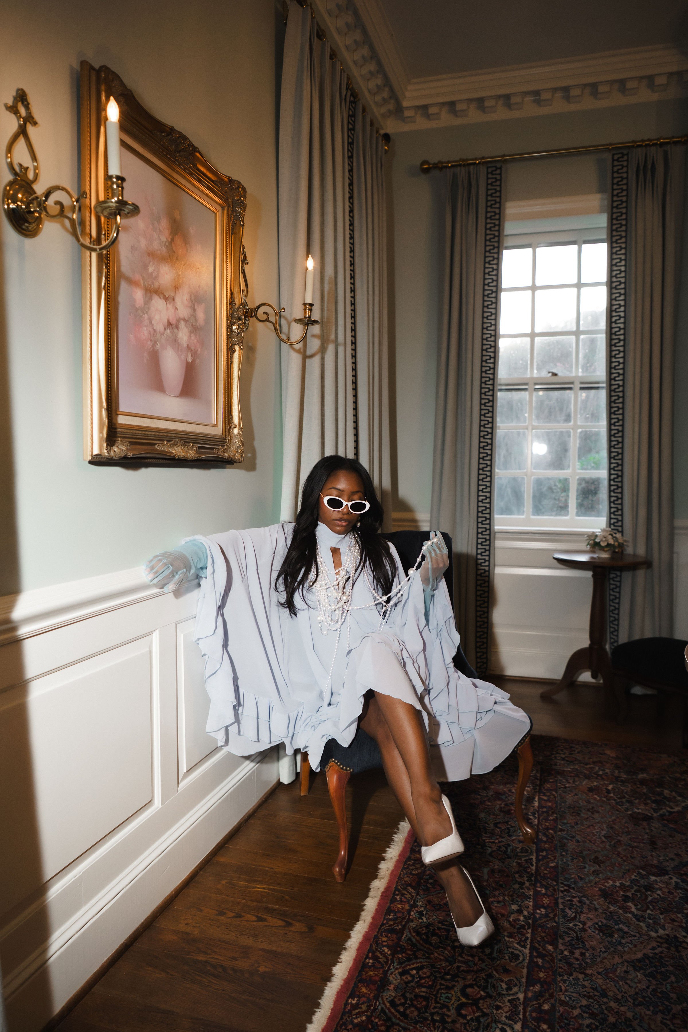 A woman sitting on a vintage chair in an elegantly decorated room with classical wall sconces, a large window with curtains, and a framed floral painting, dressed in all white, wearing white sunglasses and holding a string of pearls.