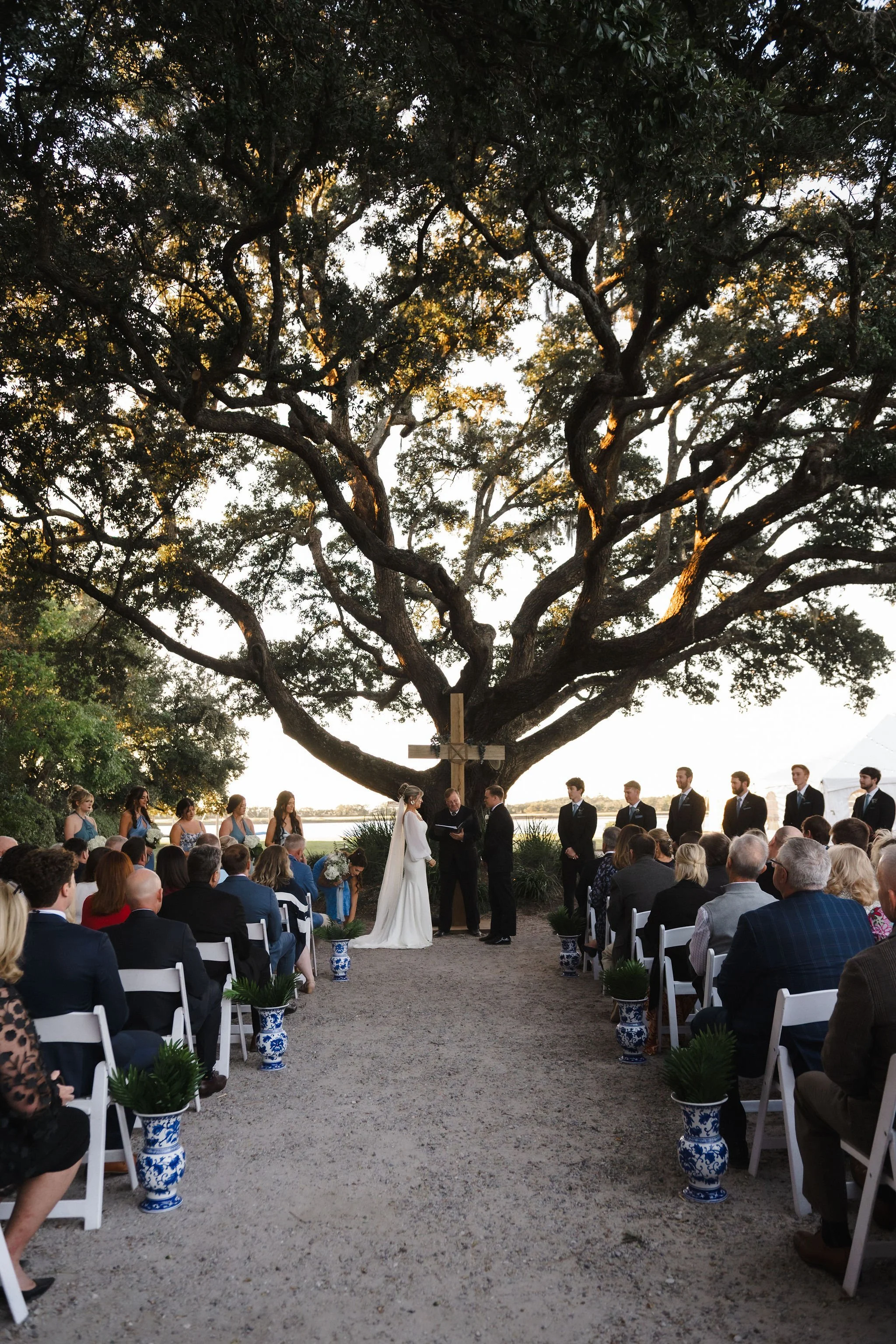 A wedding ceremony taking place outdoors under a large, sprawling tree with a cross mounted on it. The bride and groom are standing at the altar, with bridesmaids and groomsmen lined up behind them. Guests are seated on either side of the aisle, obse