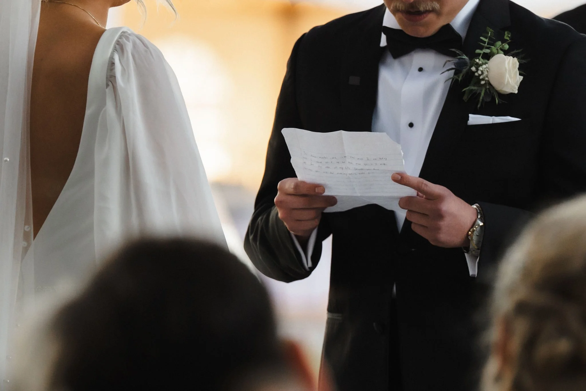 A man in a black tuxedo with a white shirt and black bow tie is reading a handwritten letter at a wedding ceremony. The man has a white boutonniere on his left lapel and is wearing a wristwatch. A woman in a white dress stands nearby, and several blu