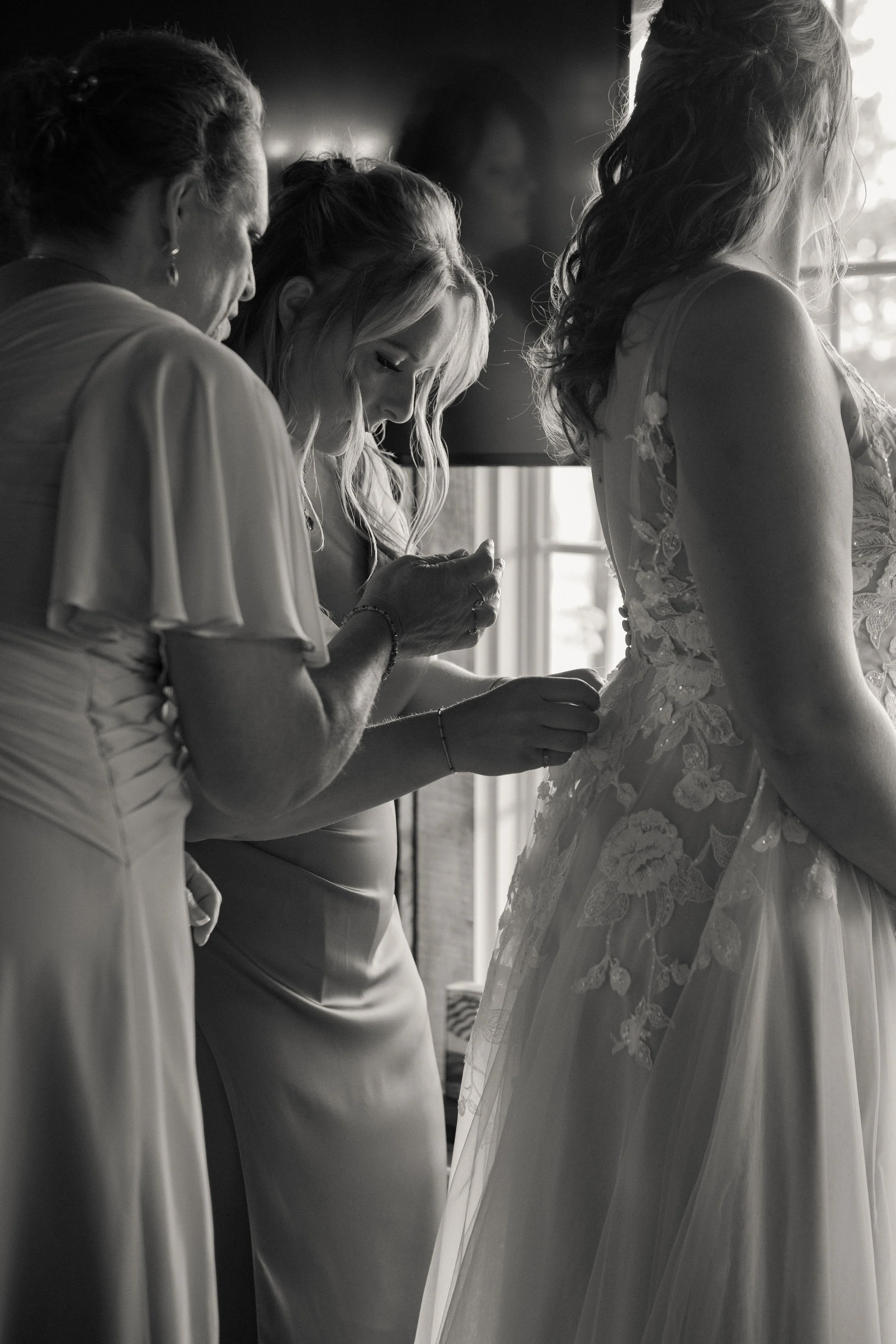 A bride in a lace wedding dress getting ready as three women help her, possibly her family members or bridesmaids, inside a room with natural light coming through a window.