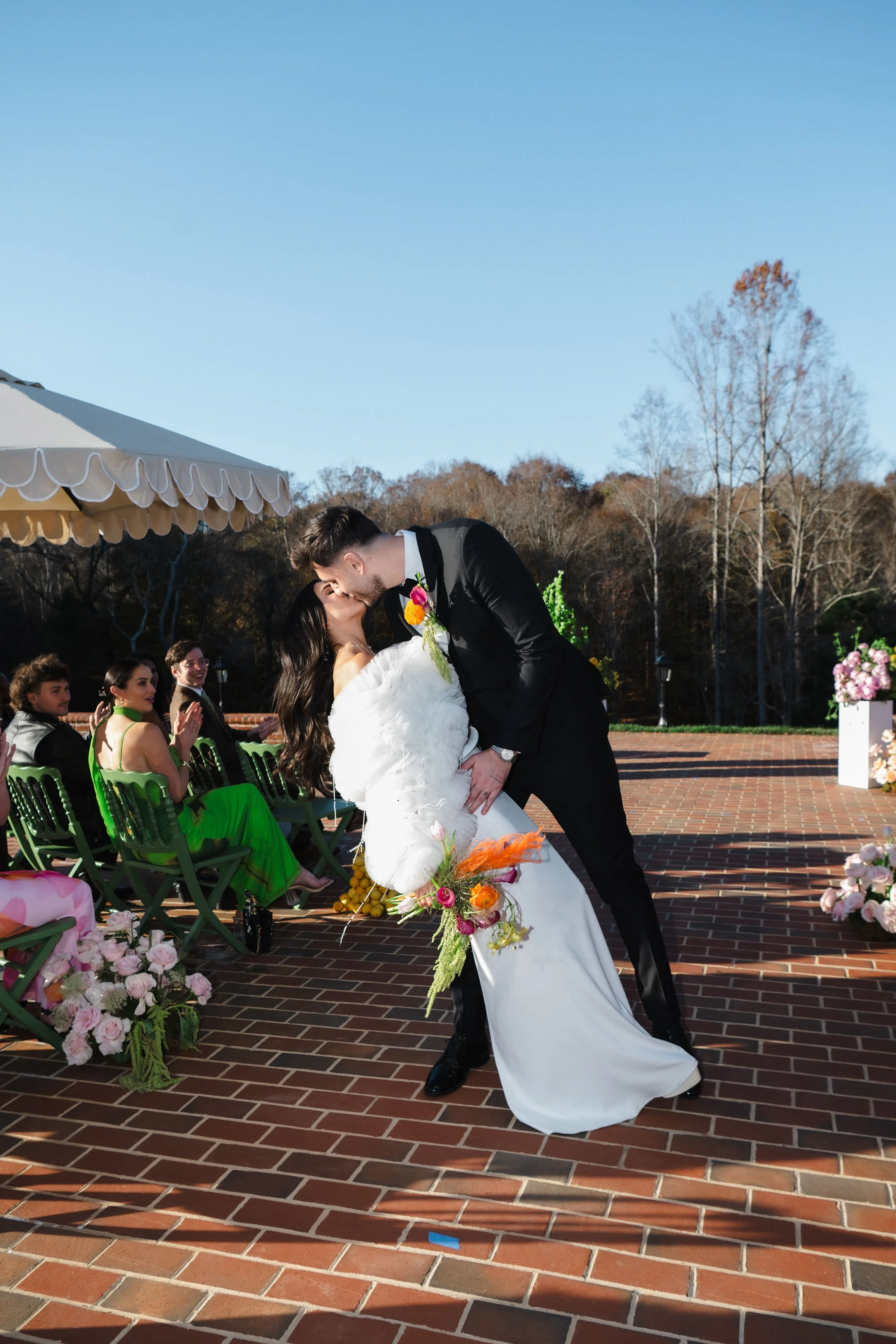 A bride and groom kiss during their outdoor wedding ceremony as guests watch.