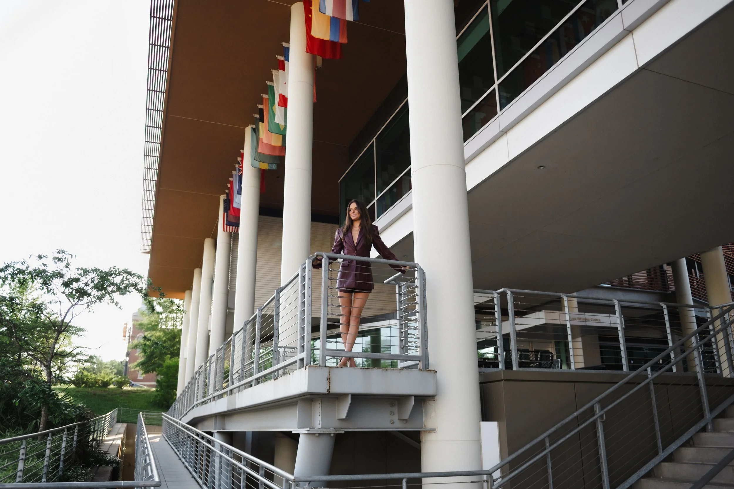 A woman standing on a balcony of a modern building with international flags hanging from poles, with trees and greenery visible nearby.