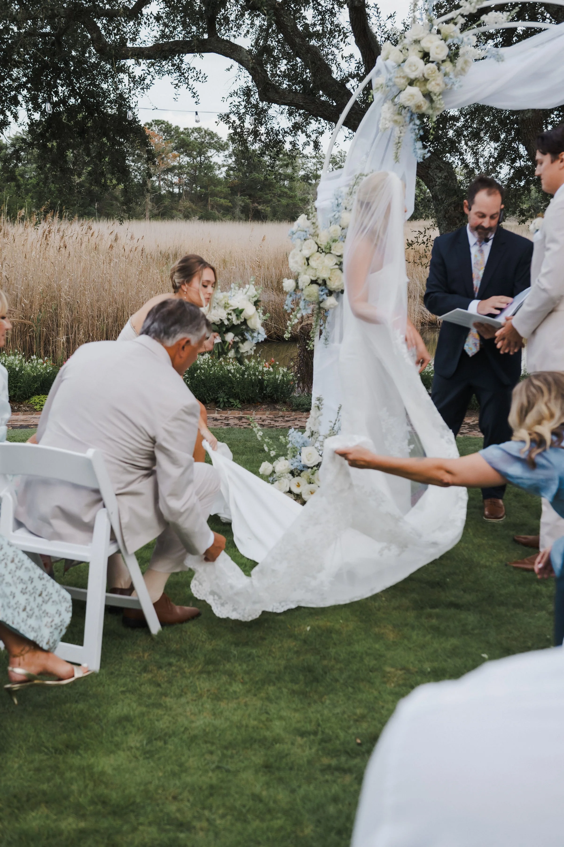 A wedding ceremony outdoors with the bride, groom, and officiant under a floral arch, with wedding party members and guests present.