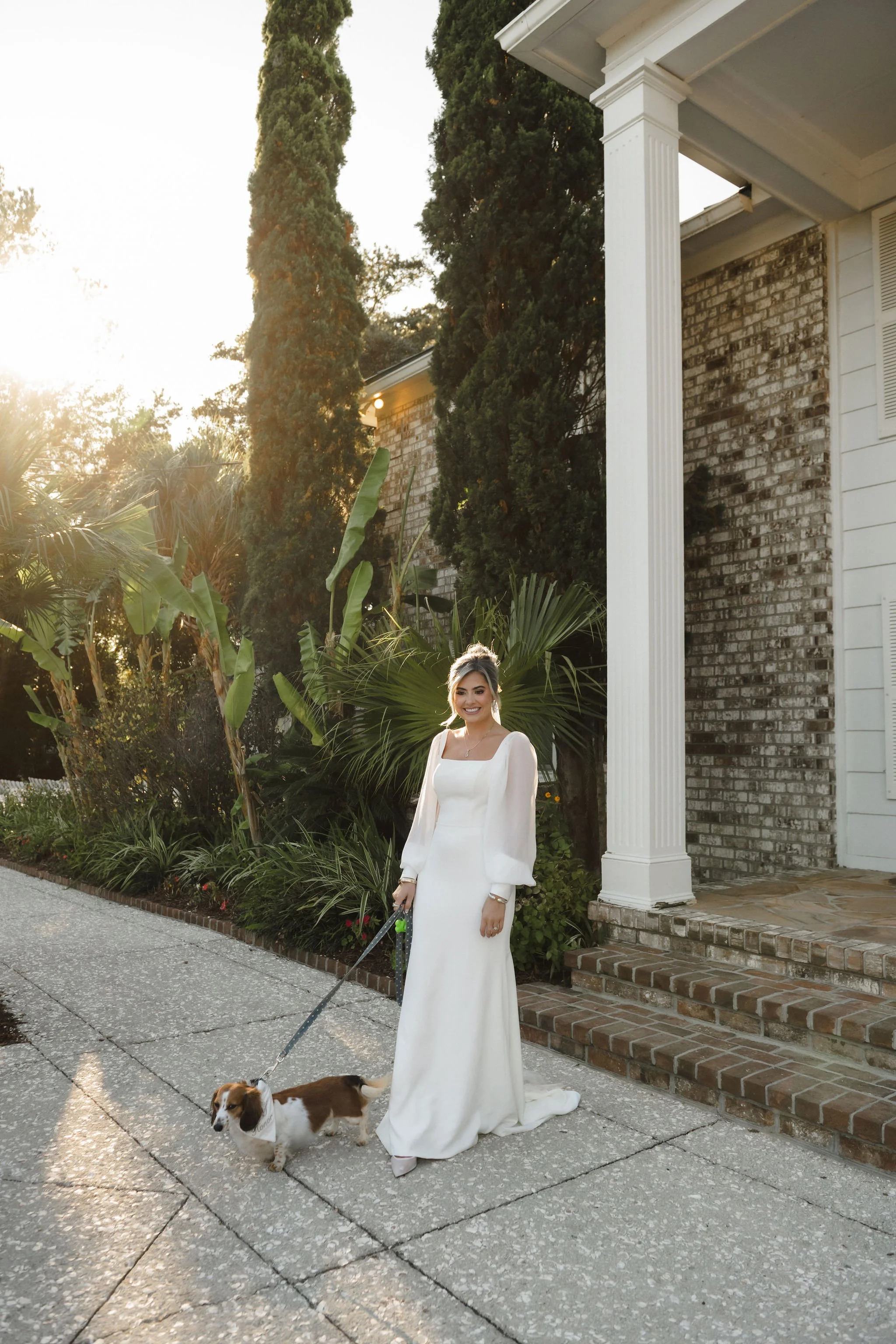 A woman in a long white dress standing on a sidewalk holding a leash attached to a small dog, with green trees and a brick house in the background, during sunset.