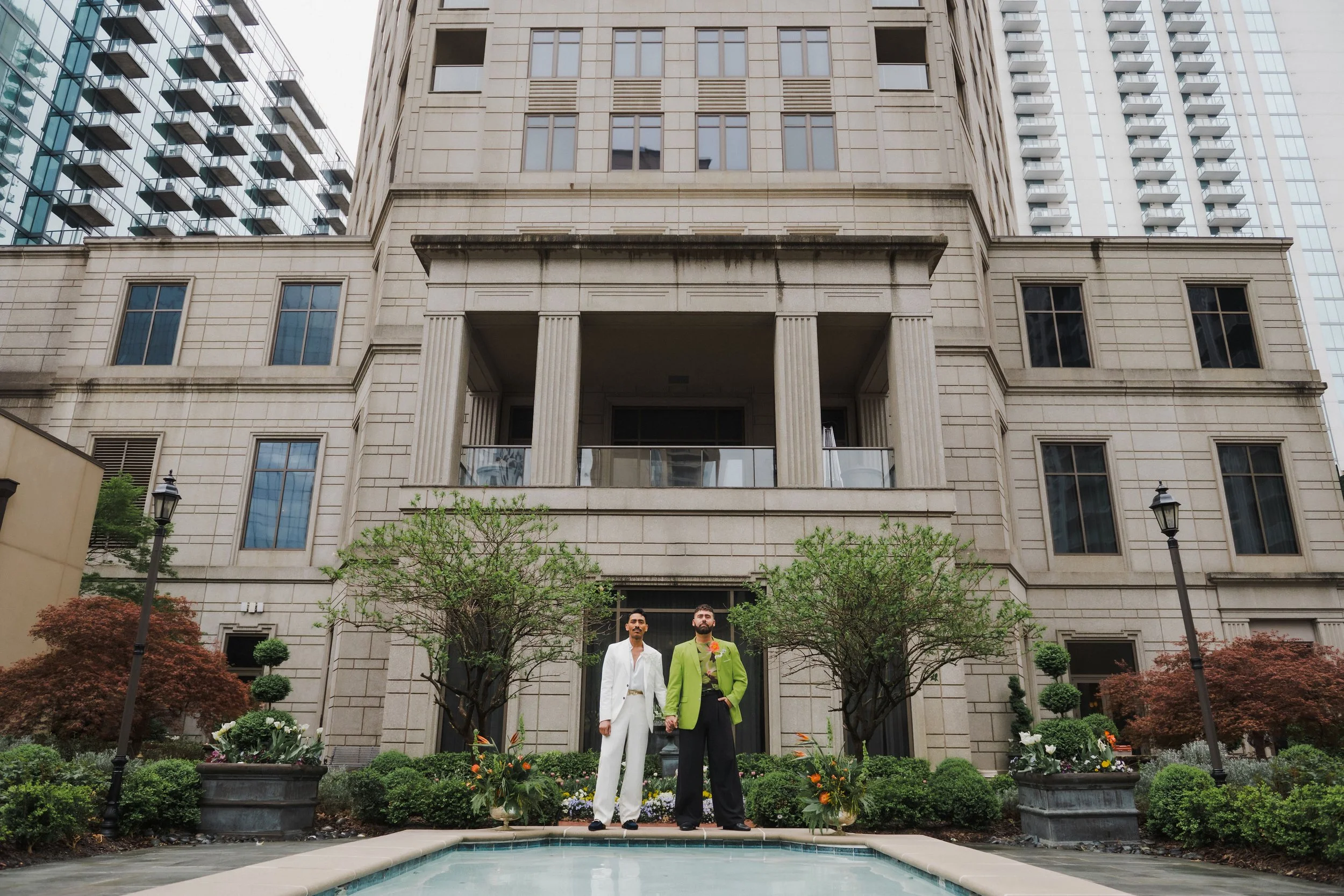 Two individuals holding hands in formal attire standing in front of a large apartment building with a garden and fountain.