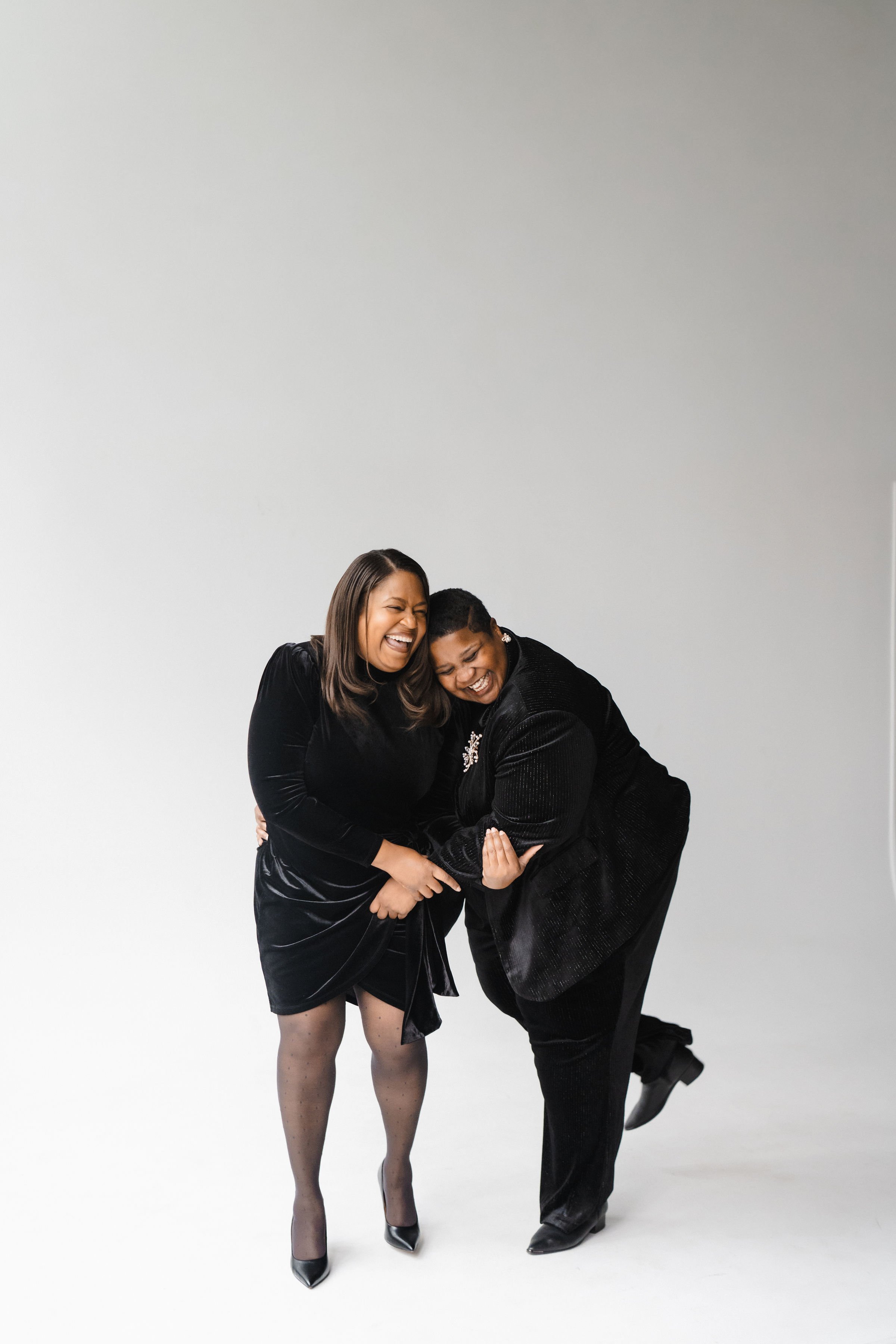 Two women in black outfits laughing and hugging each other against a plain white background.