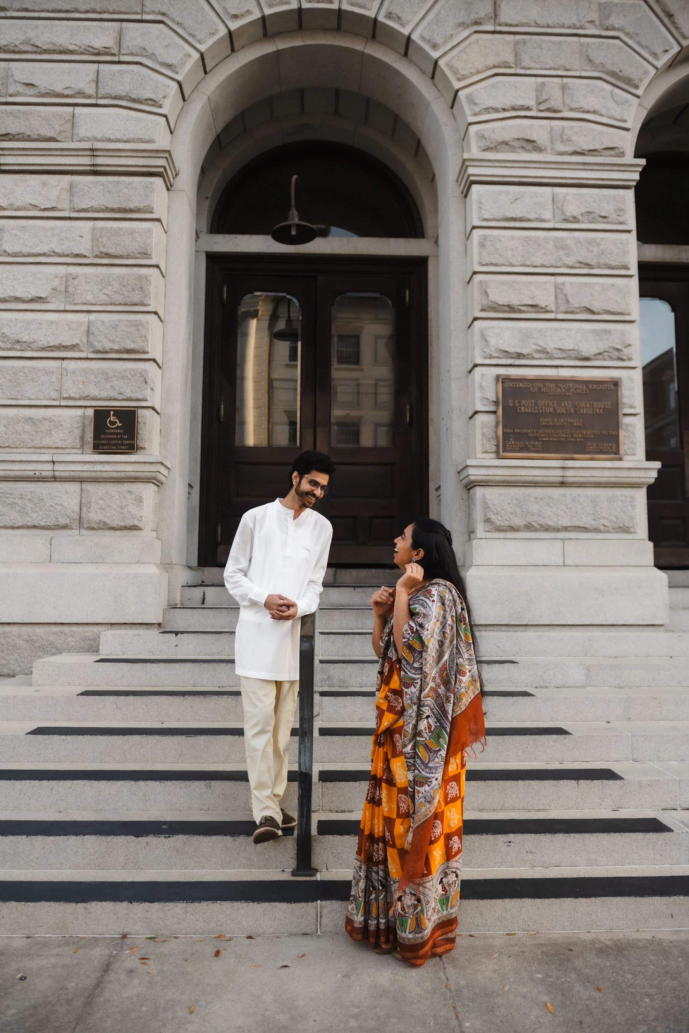 A man and woman standing on the steps of a historic building, engaged in conversation. The man wears traditional white attire, and the woman wears a colorful sari. They are smiling outside a stone building with historic plaques and a large dark woode