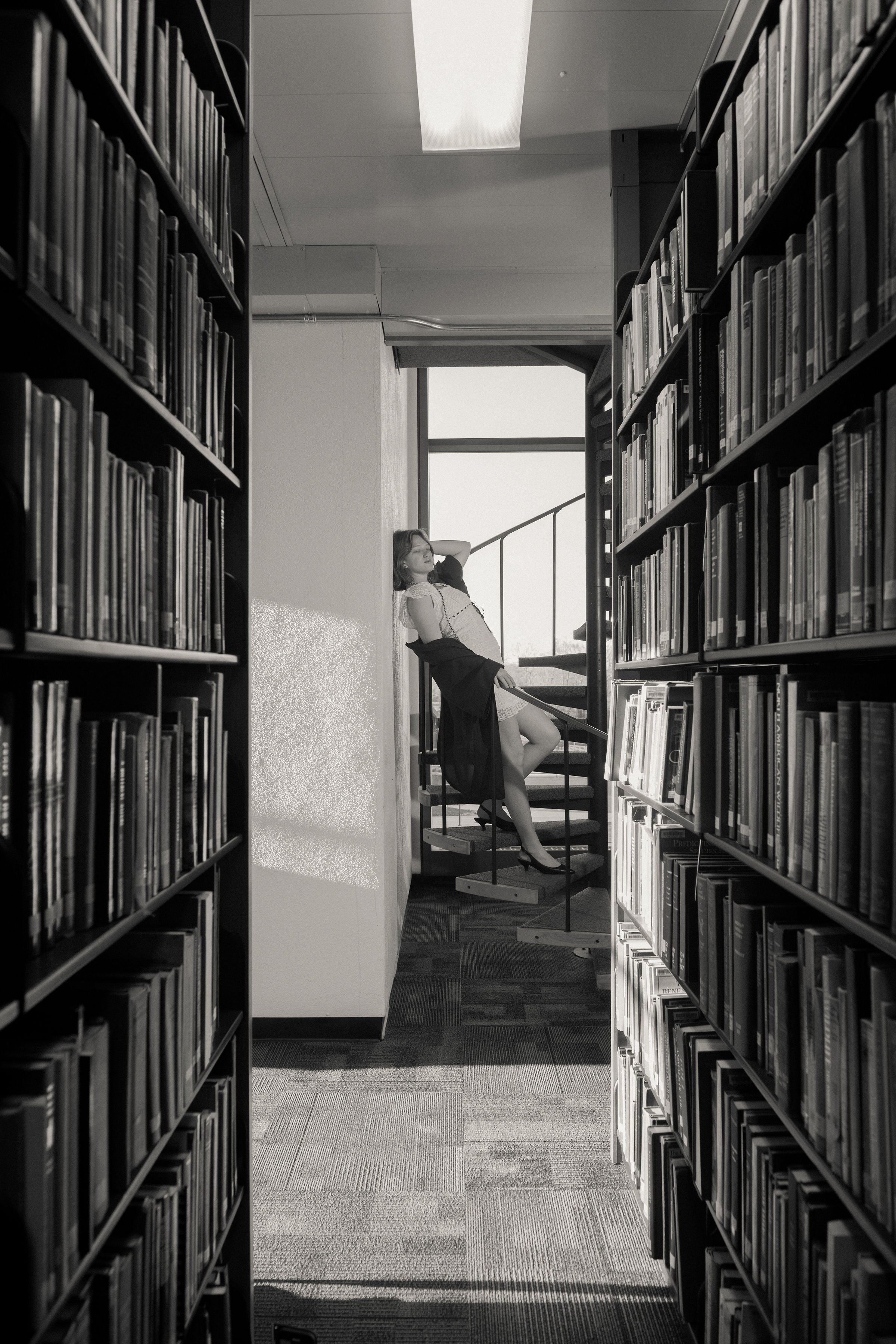A woman in high heels and a dress leaning against a wall at the top of a spiral staircase in a library, with shelves of books on either side.