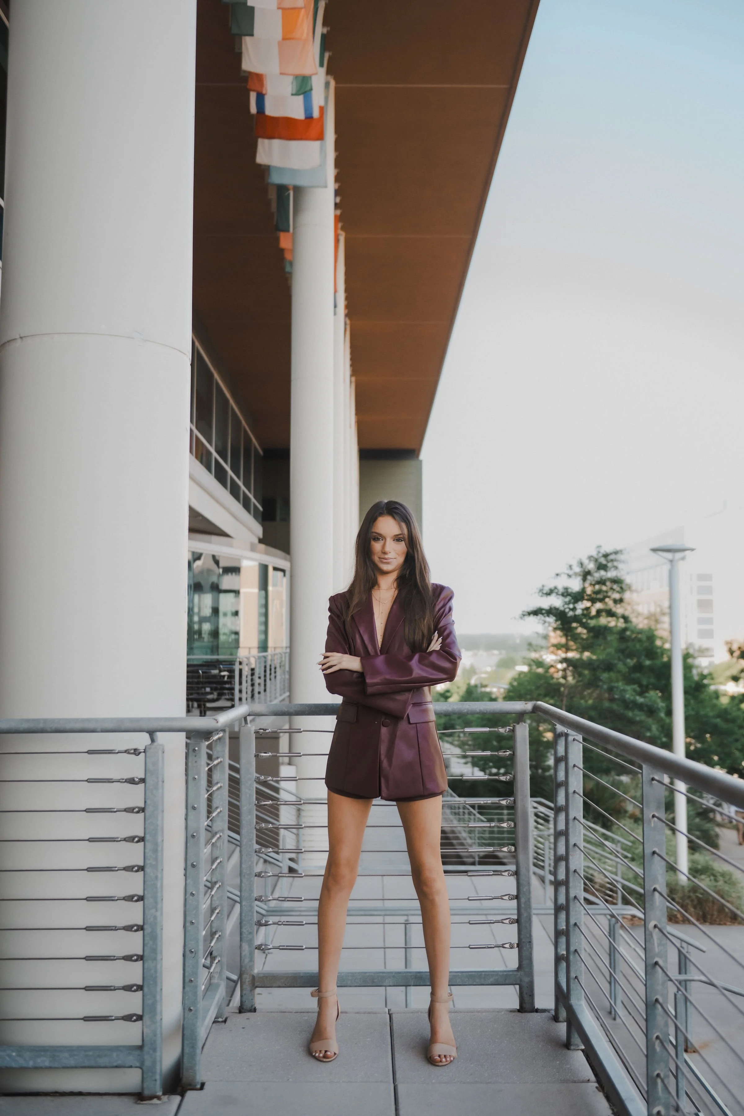 A woman standing on a balcony, crossing her arms, in a cityscape setting during daylight.