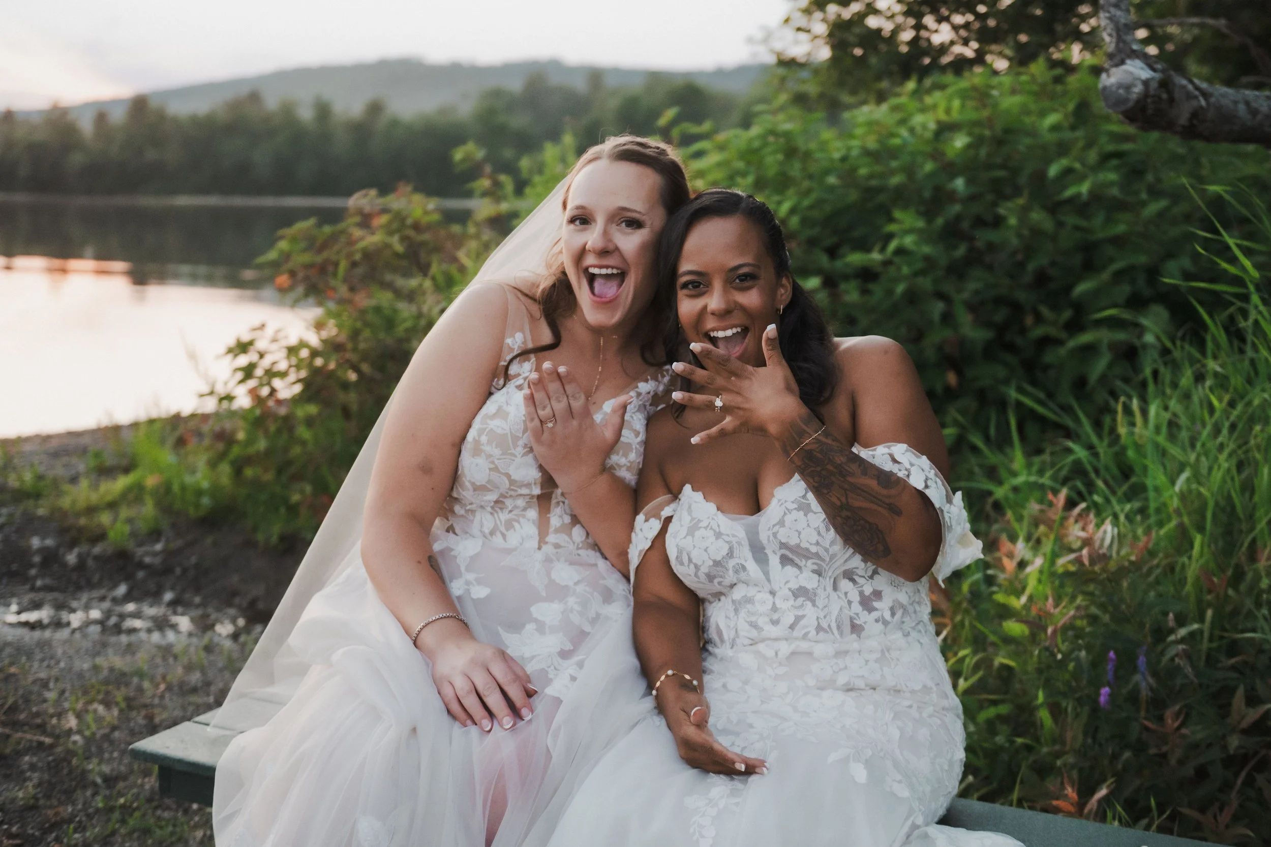 Two women in wedding dresses sitting outdoors by a river, smiling and showing off their wedding rings.