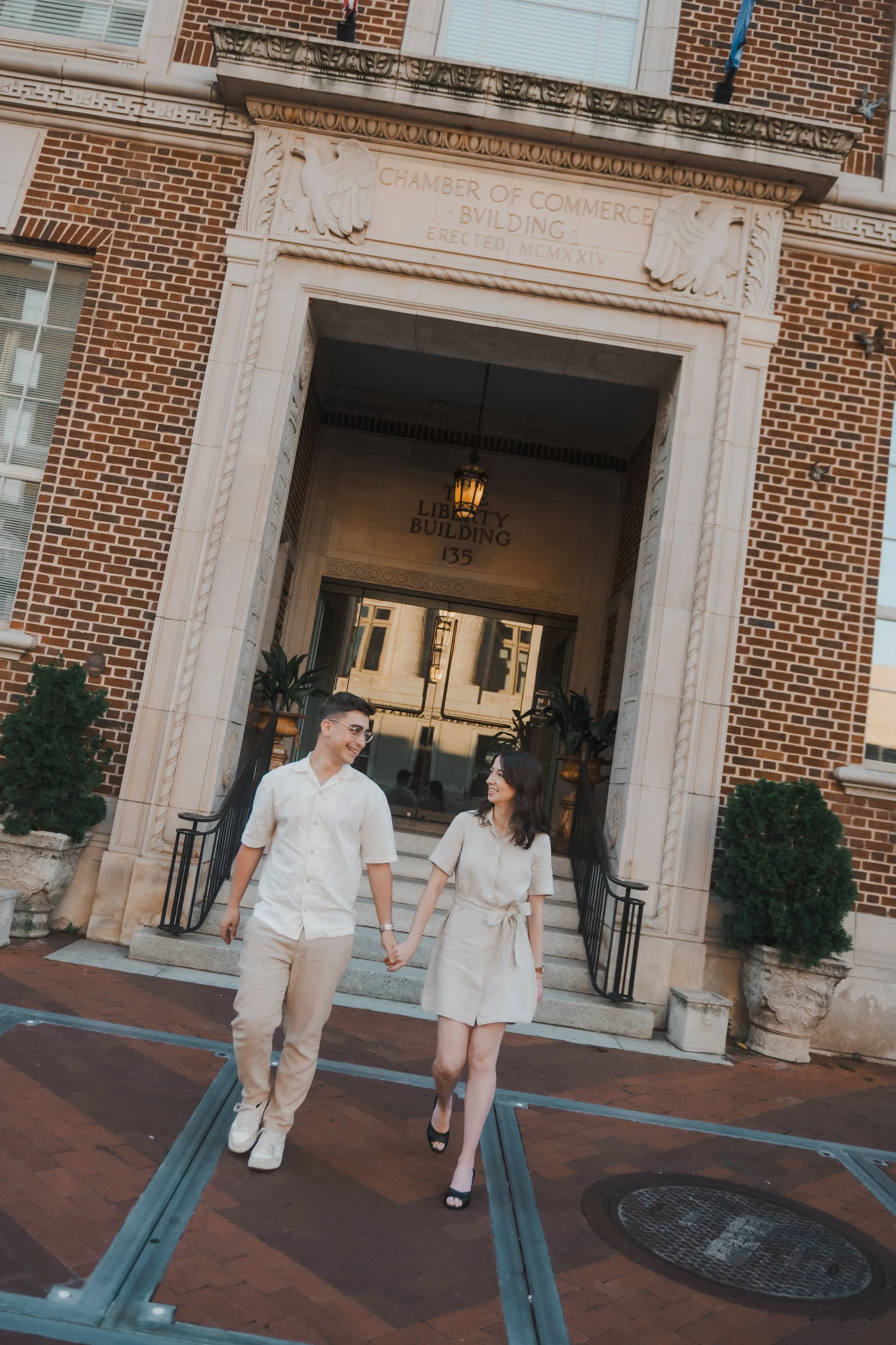 A man and woman holding hands, walking out of the Liberty Building. They are smiling at each other, dressed in light-colored clothing. The building has brick walls, decorative stonework, and potted plants at the entrance.