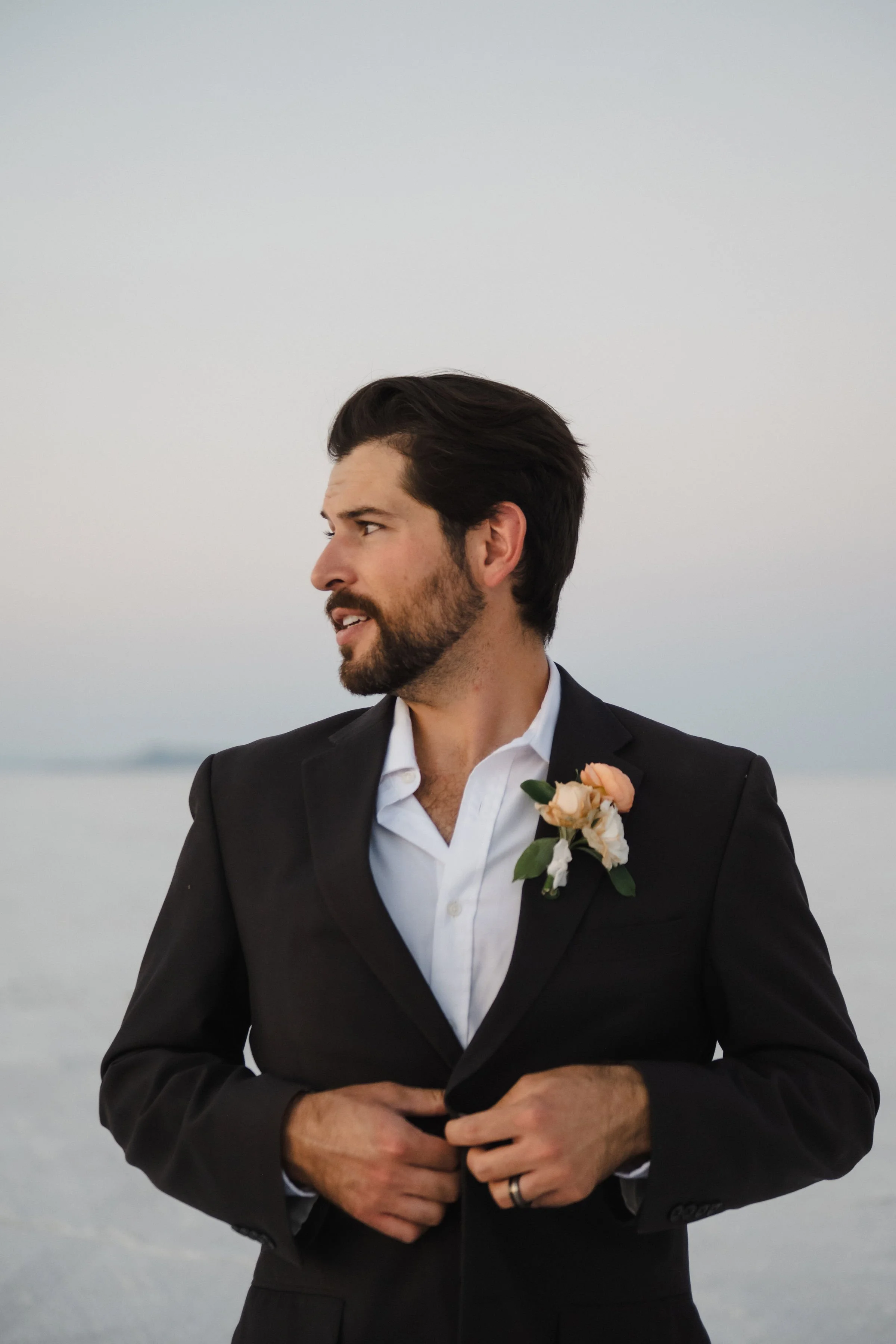A man in a black suit with a white shirt and a boutonniere stands outdoors near a body of water, looking to his left during dusk.