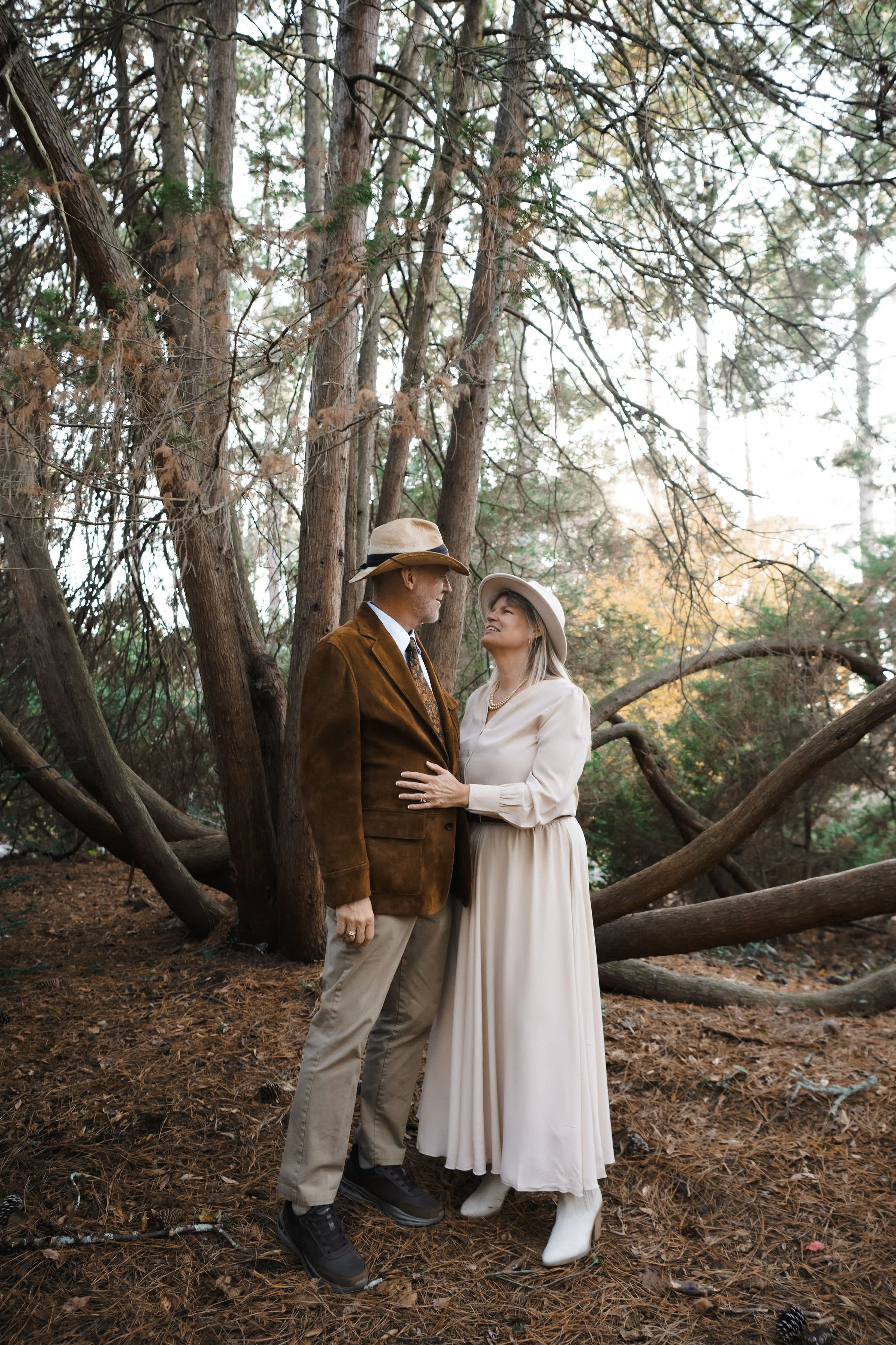 A man and woman stand close together in a wooded area during fall, gazing at each other affectionately. The man wears a brown suede jacket, beige pants, black shoes, and a hat, while the woman is dressed in a long white dress, white boots, and a wide