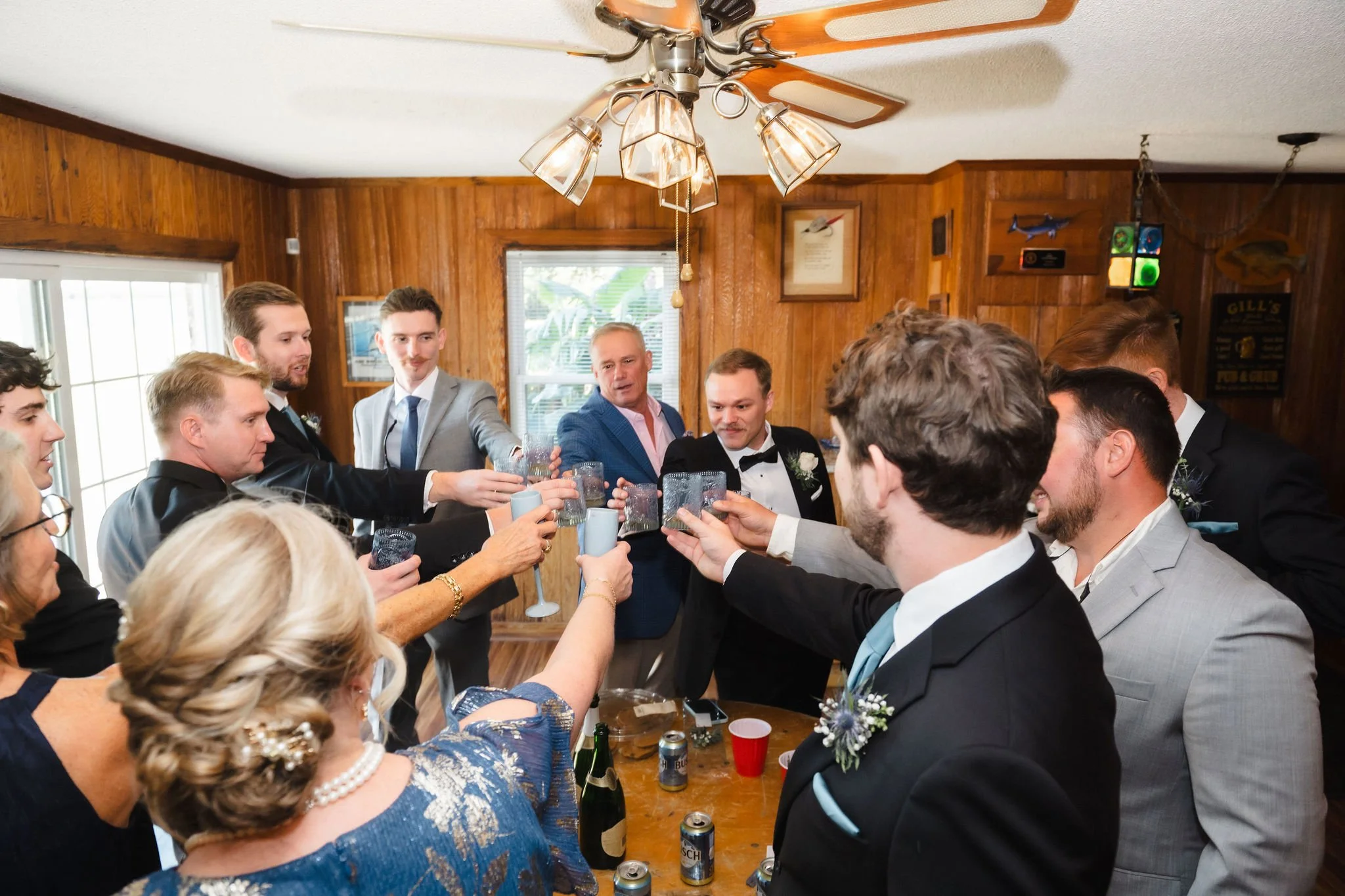 Group of people in formal attire raising glasses for a toast in a wooden-paneled room with a ceiling fan.