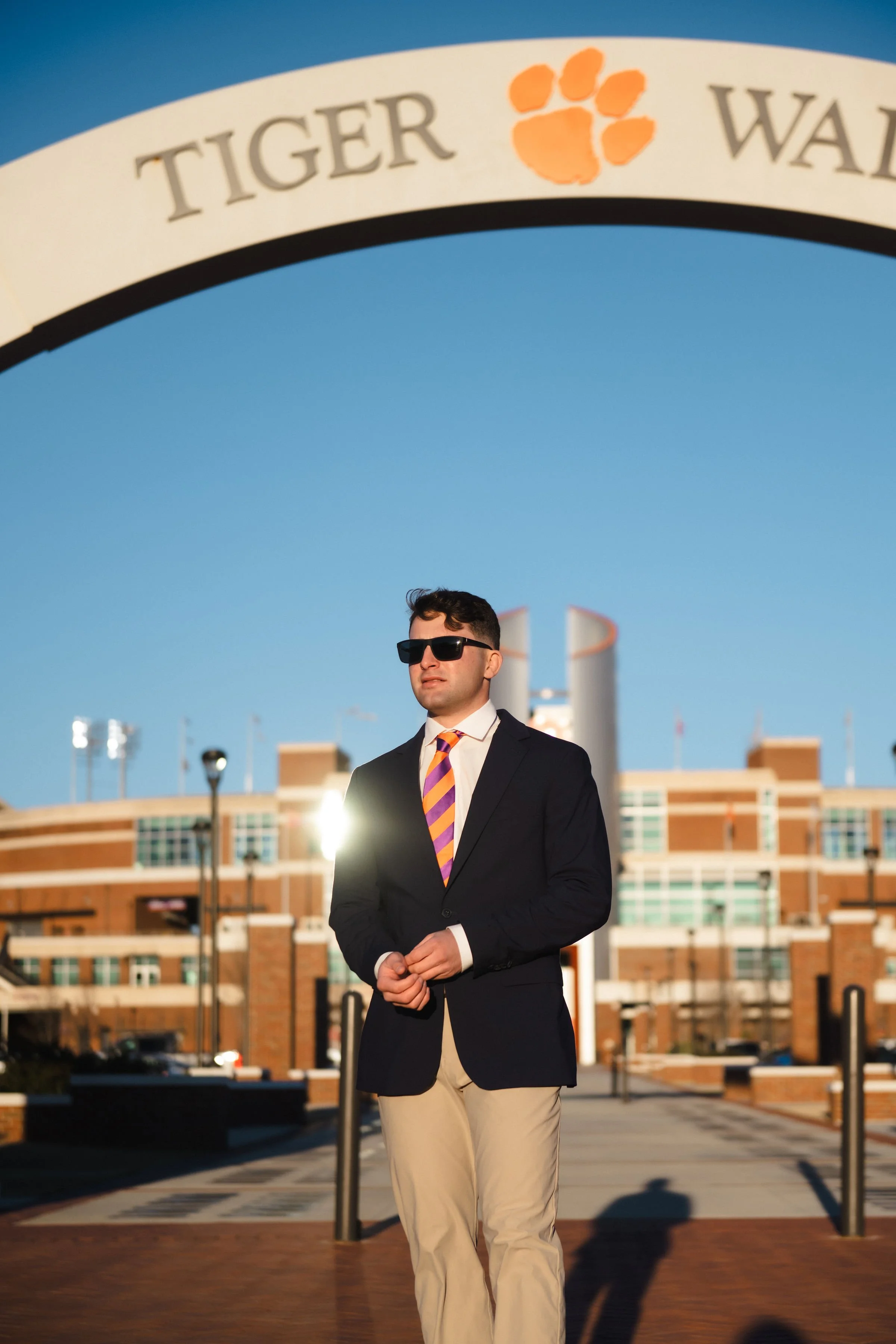 A man wearing sunglasses, a navy blazer, a colorful striped tie, and khaki pants, standing outdoors at Clemson University, with the Clemson Tigers football stadium in the background and the entrance arch overhead.