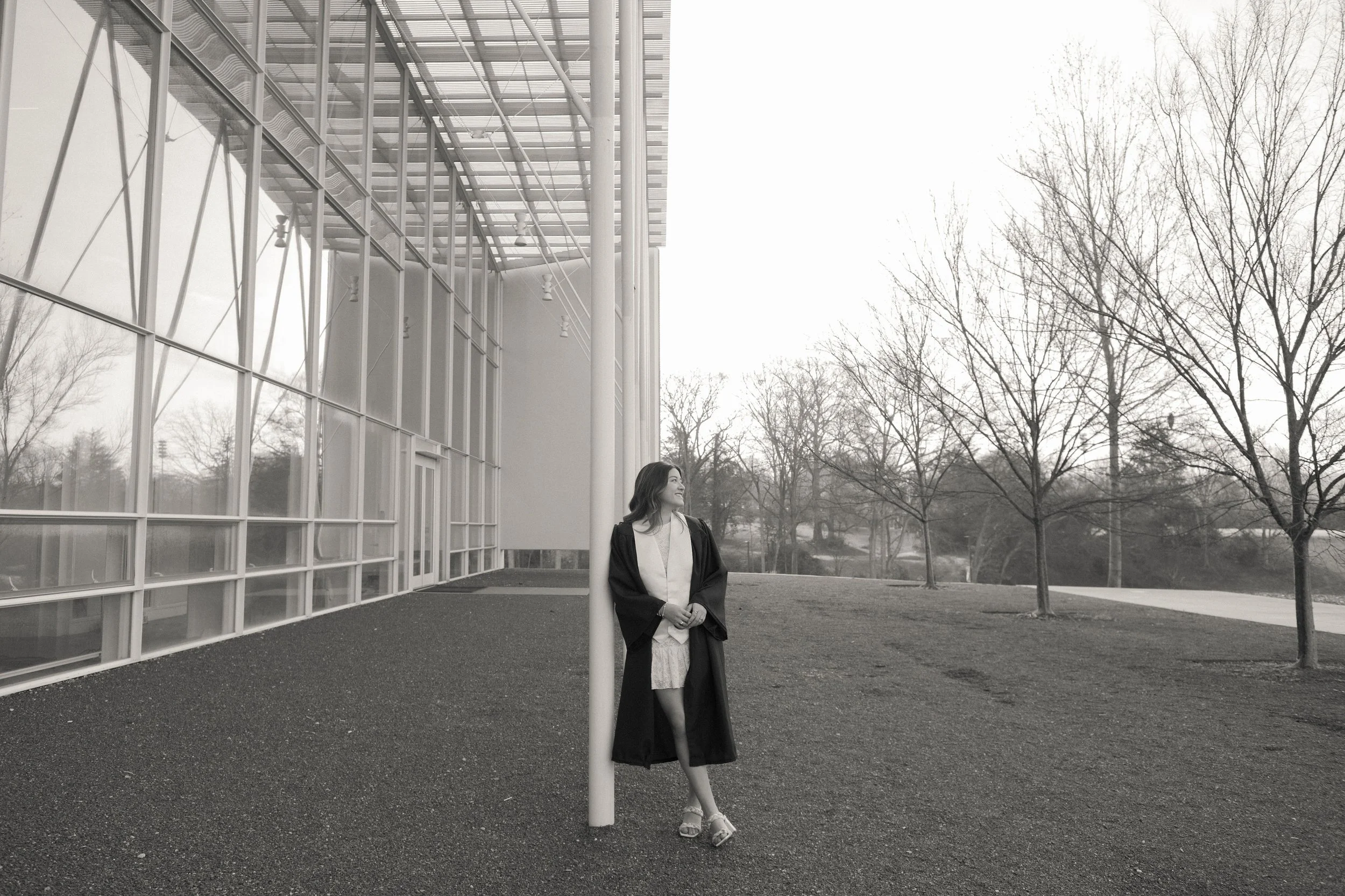 A woman in graduation attire leaning against a pole outside a modern glass building during daytime.